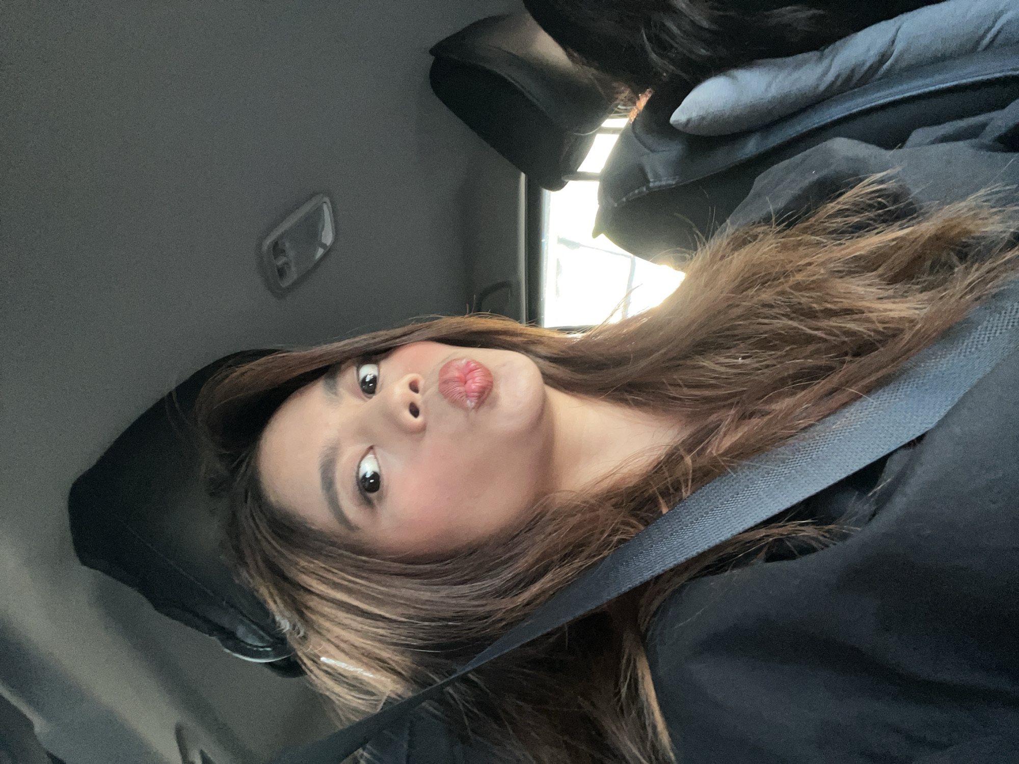 A female individual is depicted taking a selfie inside a vehicle, which is contextually located in Davao City, Philippines. The woman, estimated to be in her 20s to 30s, has long, light brown hair and is looking directly at the camera with wide eyes and pursed lips. She is wearing a dark, possibly black, garment and a dark-colored seatbelt diagonally across her torso. Makeup is visible on her face, including defined eyebrows, eye makeup, and blush. The interior of the vehicle shows a dark gray headliner with an overhead dome light fixture. Bright natural light enters the vehicle from the rear window, illuminating the scene. The top of a dark headrest is visible behind the primary subject, and a small portion of another individual's dark hair or clothing is present in the far right background.
