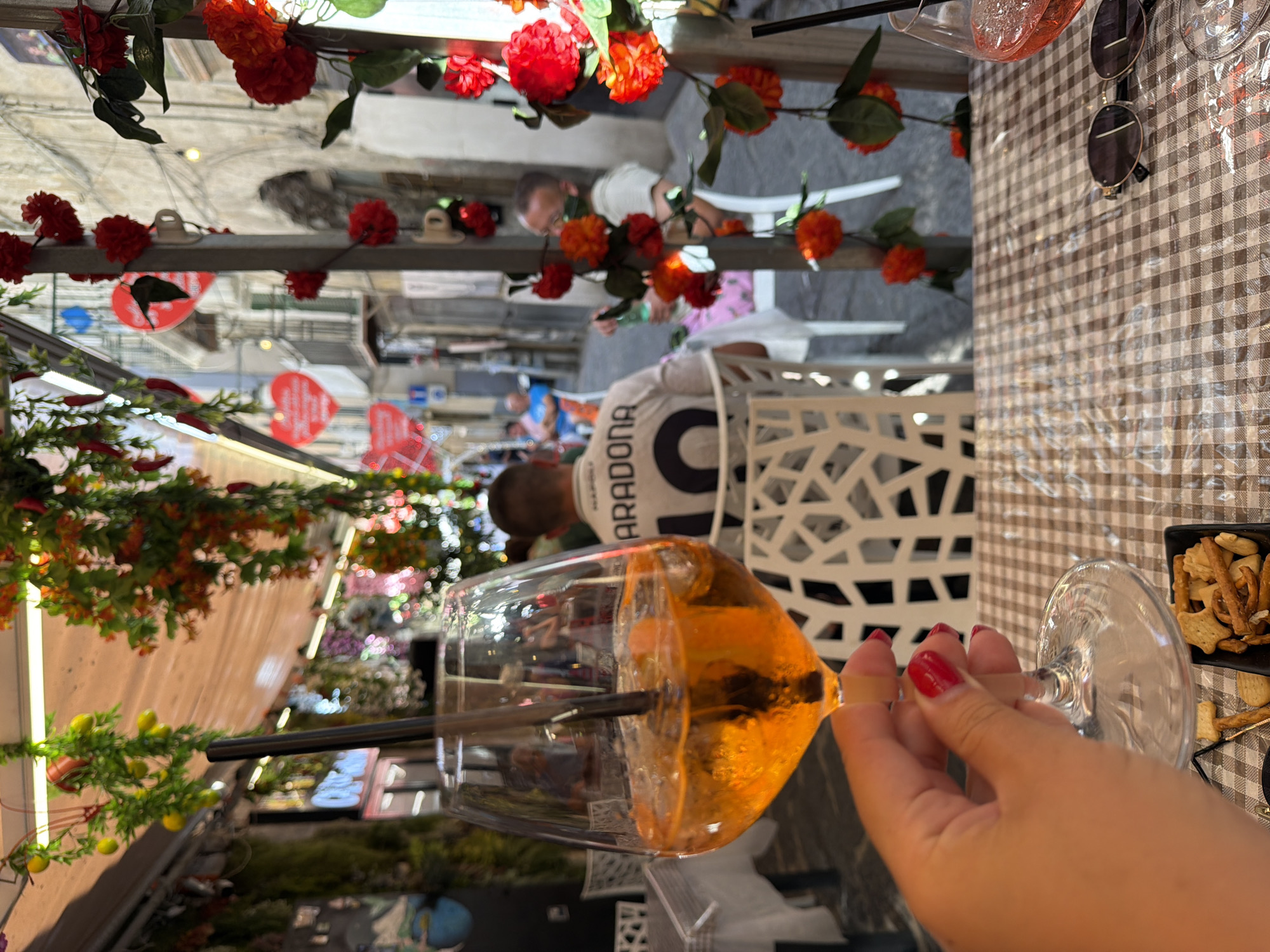The image captures a close-up of a hand with bright red nail polish holding a stemmed glass filled with an amber-colored drink, likely an aperitif. A dark straw is submerged in the beverage. The drink is set against a backdrop of a checkered tablecloth. To the right of the glass, a small black square dish contains various snacks, possibly crackers or pretzels. Partially visible in the upper right corner are a pair of sunglasses with round, dark lenses.
The background is slightly out of focus but reveals a lively outdoor scene. A person wearing a white shirt with dark lettering that appears to say "MARADONA" is seated at a table. White plastic chairs are scattered around, suggesting a restaurant or cafe setting. Decorative red and orange flowers, likely artificial, are strung along a railing or overhead structure, adding a festive touch.
Further in the background, glimpses of other people and a street or alleyway can be seen. The lighting suggests it is daytime, and the overall atmosphere appears relaxed and social, possibly indicating a leisurely afternoon or evening in Naples, Italy. The presence of drinks, snacks, and outdoor seating points to a dining or social gathering.