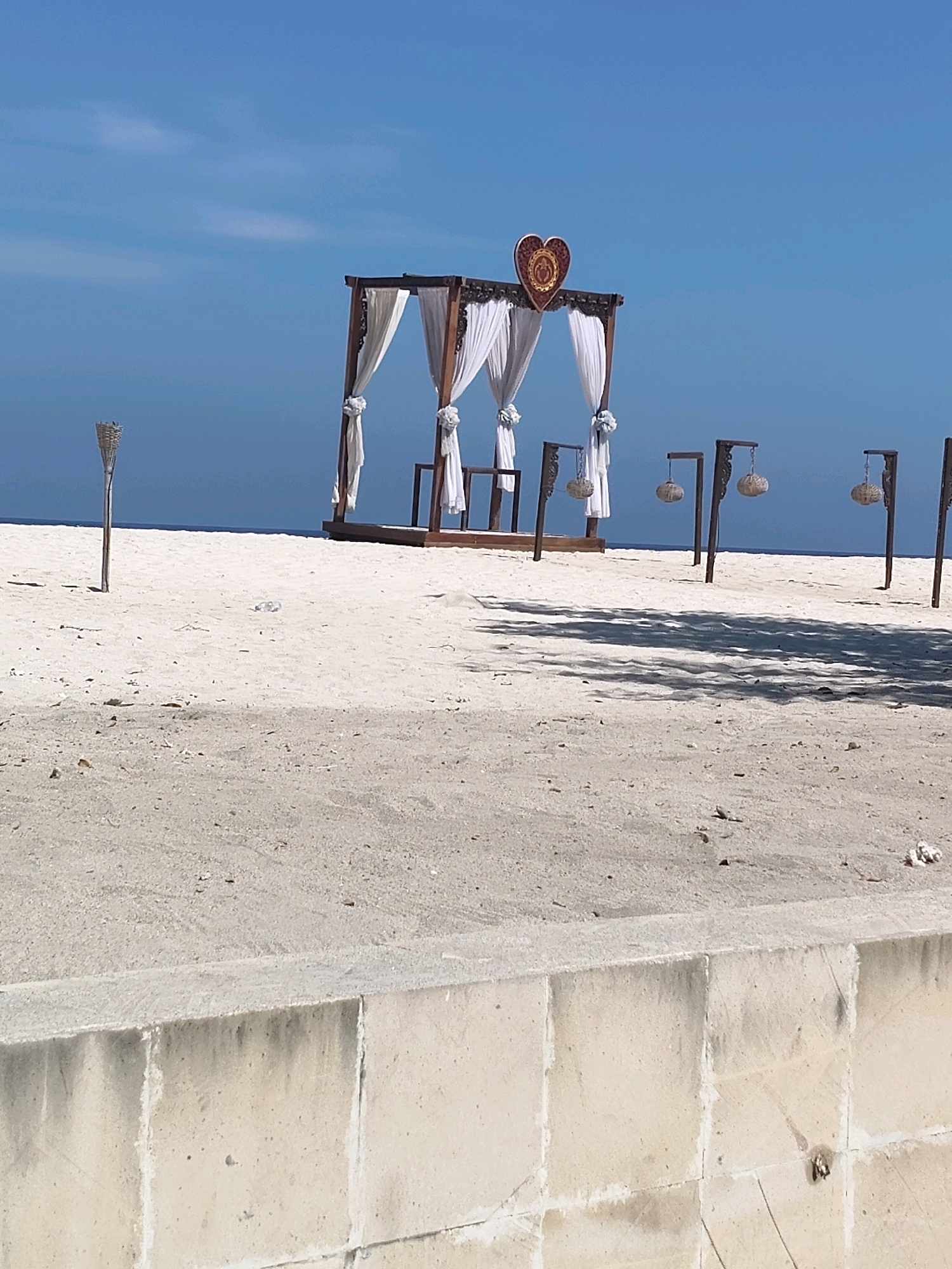 This is a medium-angle, eye-level, full shot of a decorated wedding arch on a sandy beach with a clear blue sky and ocean in the background. The arch is made of dark wood with white draping and is topped with a decorative wooden heart. There are also four decorative lanterns on wooden posts placed in a line in front of the arch. The beach is sandy with some scattered debris, and there is a concrete wall in the foreground. The location is not specified in the image or provided data.