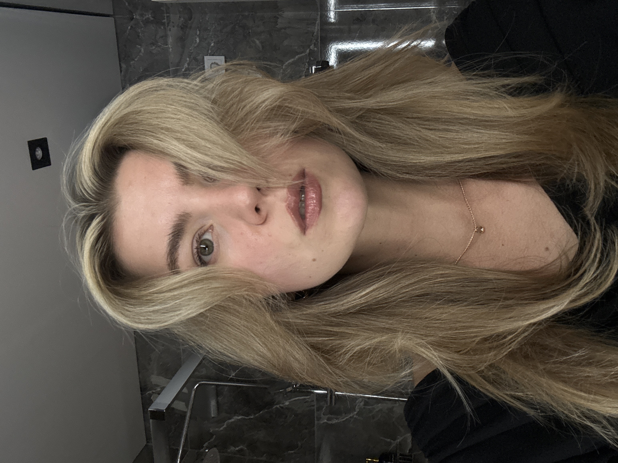 The image is a close-up shot of a person with long, blonde hair partially obscuring their face. The person's face is turned slightly to the side, revealing one green eye and a portion of their lips. They are wearing a black top, and a delicate gold necklace is visible on their neck. The background appears to be a bathroom, featuring a grey wall and a marble countertop with a chrome towel rack. The location is Unknown, Unknown.