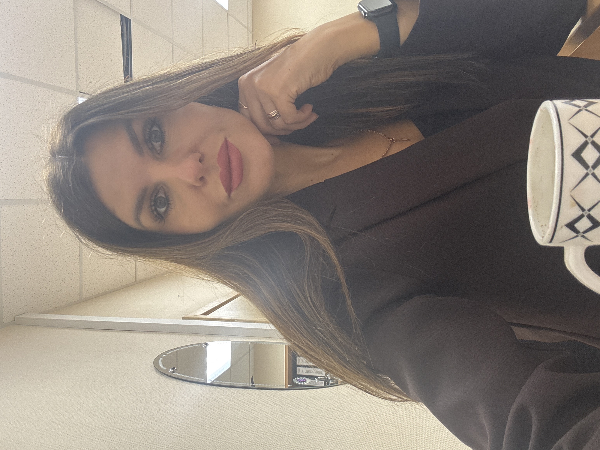A woman with long, straight, light-brown hair is posing indoors. She wears a dark-brown blazer and a black wristwatch on her left wrist. She has on a necklace and multiple rings. A mug with a black-and-white pattern sits to her right. A small, oval mirror is visible on a beige surface. The location appears to be an office setting with textured beige walls and a white ceiling with grid patterns. There is no definitive evidence to confirm the location as Chaikovsky, Russia.