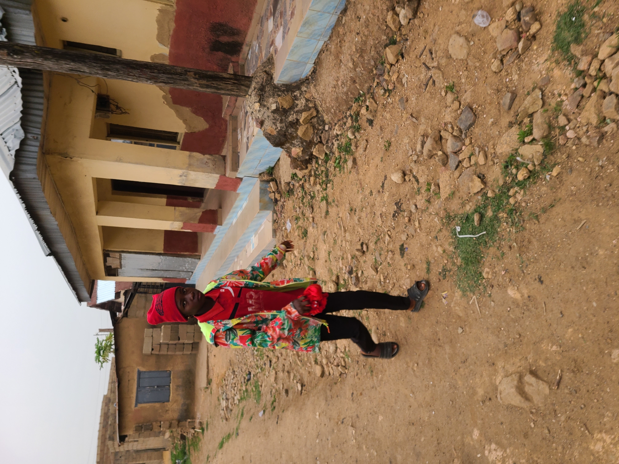 A person wearing a red head covering, a red shirt, and a floral patterned jacket stands on a dirt road in Jos, Nigeria. The ground is covered with dirt and rocks. Behind the person, there are buildings of varying heights and construction styles. The buildings are painted in shades of yellow, red, and brown. The sky is overcast.