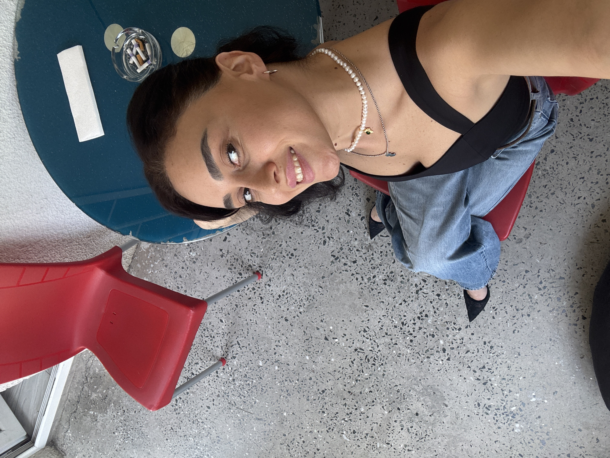 The image depicts a woman sitting on a red chair at a table, likely in an outdoor seating area of a cafe or restaurant in Tbilisi, Georgia. The floor is a speckled concrete material. A round, dark blue table is visible, with an ashtray containing cigarette butts, a small white paper object, and a circular pattern on its surface. The woman has dark hair pulled back, is wearing a black top and blue jeans, and is looking up at the camera with a smile. She has a pearl necklace with a pendant and a thin silver chain with a cross pendant. Her feet are visible, wearing black pointed-toe shoes. A red chair is prominently featured in the foreground. The lighting suggests it is daytime.