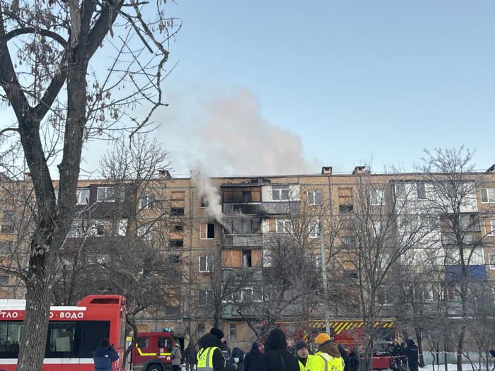 A multi-story brick residential building with extensive fire damage is observed under a clear blue sky in Unknown, Unknown. Significant charring and smoke emissions originate from a fourth-floor balcony/window area and a section of the top floor. Plumes of light grey smoke rise into the sky. The building's facade is light brown brick, with several broken or boarded-up windows on the upper floors. Bare deciduous trees with visible branches are positioned in the foreground and mid-ground, suggesting a cold season, and patches of snow are visible on the ground.
In the foreground, multiple individuals are gathered. Several are dressed in winter clothing and wear high-visibility yellow/green vests, indicative of emergency personnel or officials. One individual on the far left, near a large red vehicle, is holding a camera. The red vehicle, partially visible on the left, bears white Cyrillic text "ТЛО - ВОДА" on its side. A red fire truck with yellow stripes and the number "18" is partially visible in the mid-ground, and another red emergency vehicle is partially visible on the far right. A red and white striped barrier tape runs along the ground, demarcating a perimeter. The scene suggests an active or recently concluded fire incident with emergency response present.