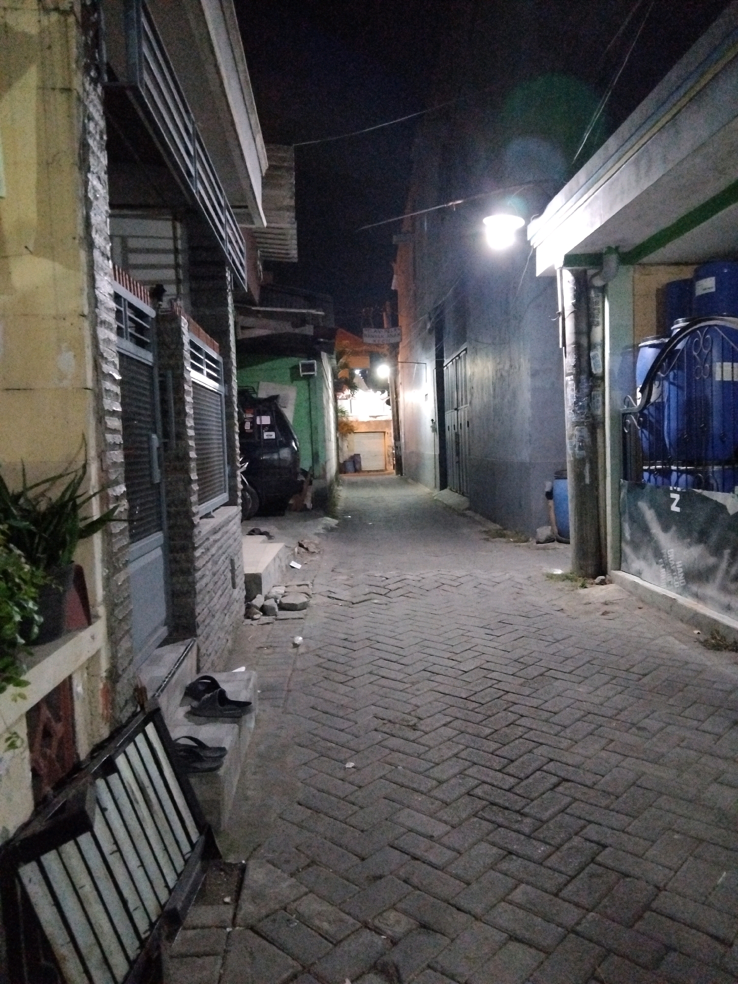 The image depicts a narrow, paved alleyway at night in Kepuh Kiriman, Indonesia. The alley is flanked by buildings on both sides. On the left, a building has a yellow-toned wall and a stone-clad entrance with a metal gate. A potted plant sits on a ledge in front of this building. On the right, a building has a gray-toned wall with a white and green striped overhang. A metal gate with decorative scrollwork is visible, behind which are two large blue water barrels. A street light illuminates the alley from the right side. The alleyway floor is paved with dark rectangular bricks laid in a herringbone pattern. In the foreground on the left, there is a metal grate structure. On a concrete step in front of the building on the left, a pair of dark-colored flip-flops are placed. Further down the alley, a dark-colored vehicle, possibly an SUV, is parked. The overall impression is of a residential or commercial back alley at night.