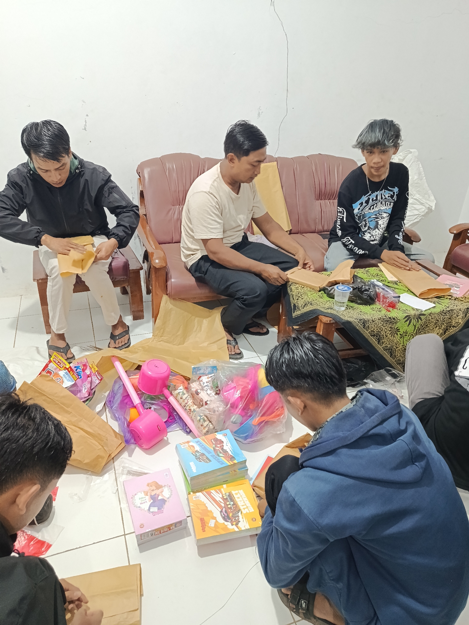 In Pocol, Indonesia, four individuals are gathered in a room with white walls. Two men are seated on a brown leather sofa, while another two men are on the floor. One man on the sofa, wearing a black t-shirt with a graphic design, is looking down at a brown paper envelope in his hands. The man next to him, in a beige t-shirt, is also looking down, seemingly engaged in a similar activity. On the floor, a man in a black jacket is meticulously folding a brown paper envelope. Another man, wearing a blue hoodie, is crouched on the floor, also interacting with brown paper bags and packages.
In the foreground, a collection of items is spread across the floor, including several children's books with colorful covers, pink toys such as a water gun and a spinning top, and various plastic bags containing what appear to be gifts or party favors. The overall scene suggests a group activity, possibly the preparation of gift bags or care packages, within what appears to be a residential setting. The lighting suggests it is nighttime.