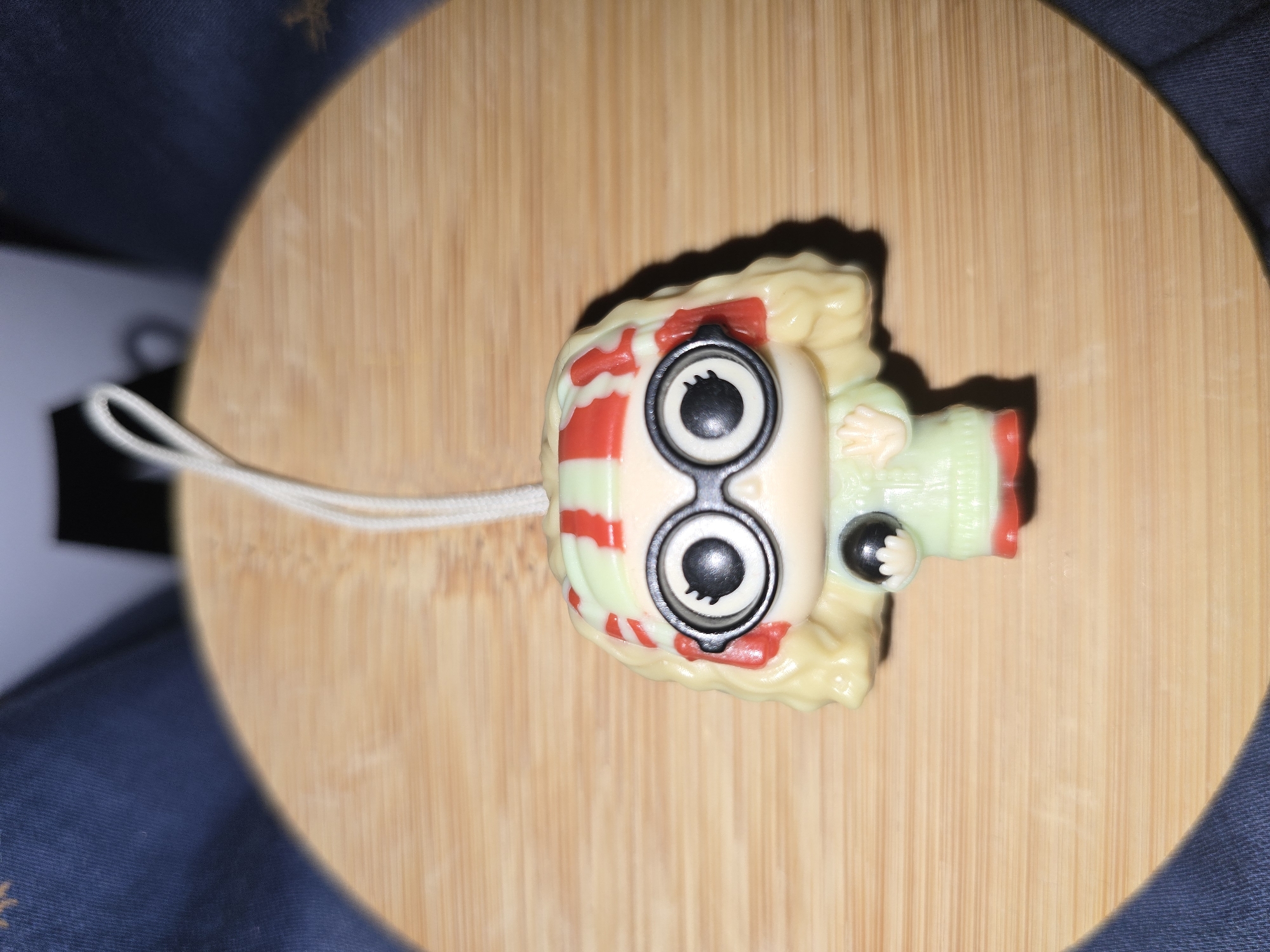 A Funko Pop keychain of a blonde-haired character wearing a light green sweater with red stripes on the collar and cuffs, red shoes, and large black-rimmed glasses with white and black irises is centered on a round, light brown wooden surface. The character has a black sphere in its left hand and a white string loop attached to the top of its head. The background consists of dark blue fabric with some texture. This image was taken in Petropavlivska Borshchahivka, Ukraine.