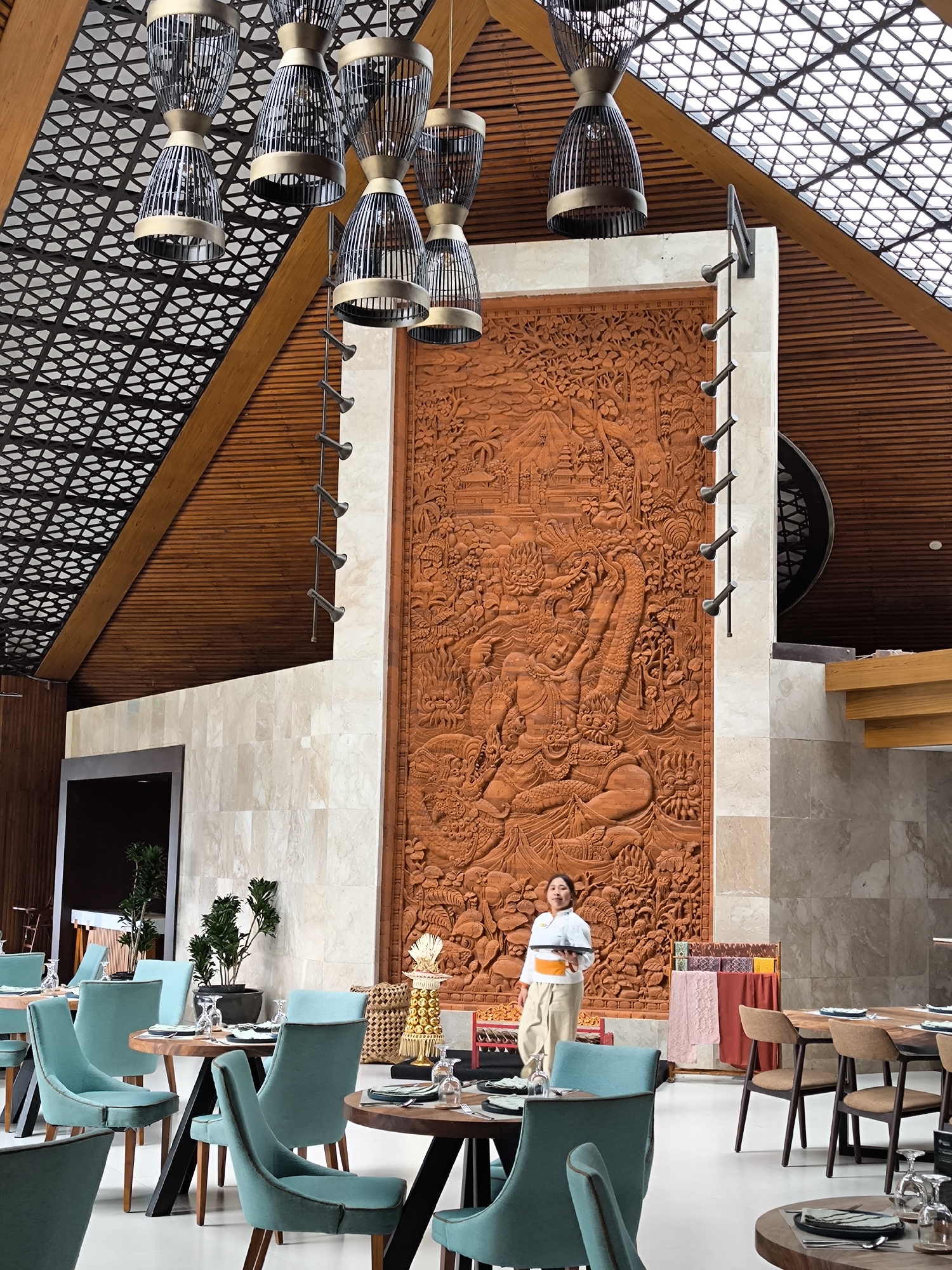 The image depicts the interior of a dining establishment in Wanagiri, Indonesia. Dominating the scene is a large, intricate terracotta-colored relief carving on a wall. In front of this carving stands an Asian woman in a white top and light-colored pants, wearing an orange sash around her waist. She is holding a white tray.
The space is furnished with tables and chairs. The chairs are upholstered in a soft blue hue, and the tables are dark wood with minimalist metal frames. The lighting is provided by a series of hanging metal lamps with a conical design. The ceiling is geometrically patterned, with exposed wood beams. Plants are interspersed throughout the dining area. The floor is white. The scene suggests a fine dining atmosphere with a strong cultural aesthetic influence.