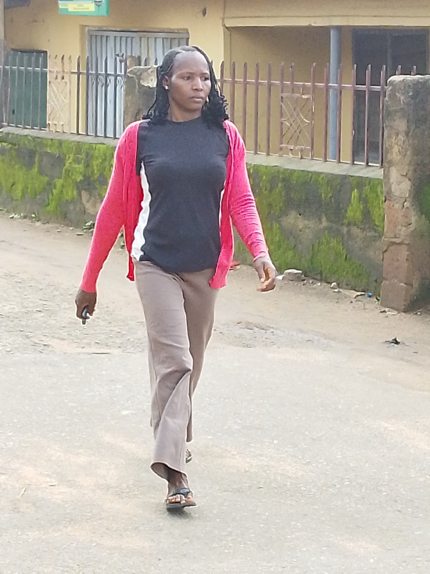 A medium shot shows a woman walking on a paved road in what appears to be a city. She is facing to her right and is wearing a pink cardigan over a black and white shirt, with tan pants and dark sandals. She is holding a dark-colored smartphone in her left hand. The background features a low wall covered in moss and green foliage, with a metal fence behind it. Beyond the fence, a building with a pale yellow facade and metal security bars is visible. The environment appears to be outdoors and possibly in a residential or commercial area of a city.