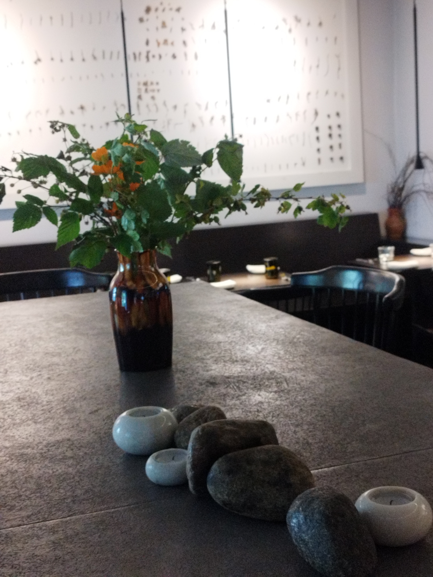 The image depicts a dimly lit restaurant interior. In the foreground, a dark, textured table is partially visible. Several smooth, dark stones are arranged in a line on the table, accompanied by two small white candle holders, one with a lit candle and another unlit. Further back on the table, a tall, dark brown glass vase holds a bouquet of green leaves and small orange flowers. Behind the table, dark chairs are visible, and in the background, a white wall features a decorative arrangement of small, dark vertical elements. The overall impression is one of a sophisticated and possibly intimate dining setting. There is no information to suggest the location is Saint Petersburg, Russia.
