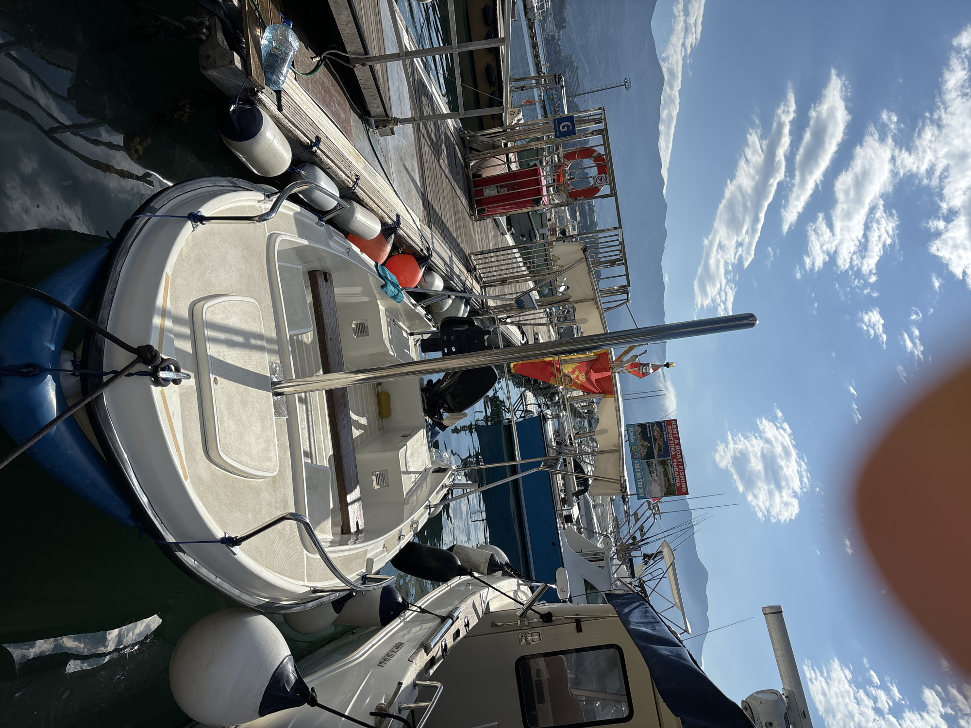 The image depicts a scene at a marina in Budva, Montenegro. A small white boat with blue fenders is docked alongside a wooden pier. A life preserver and other marina equipment are visible on the pier. Further back, other boats are docked. In the background, mountains rise against a partly cloudy sky. A sign advertises boat rentals and fishing trips and includes the phone number +382 69 730 361.
