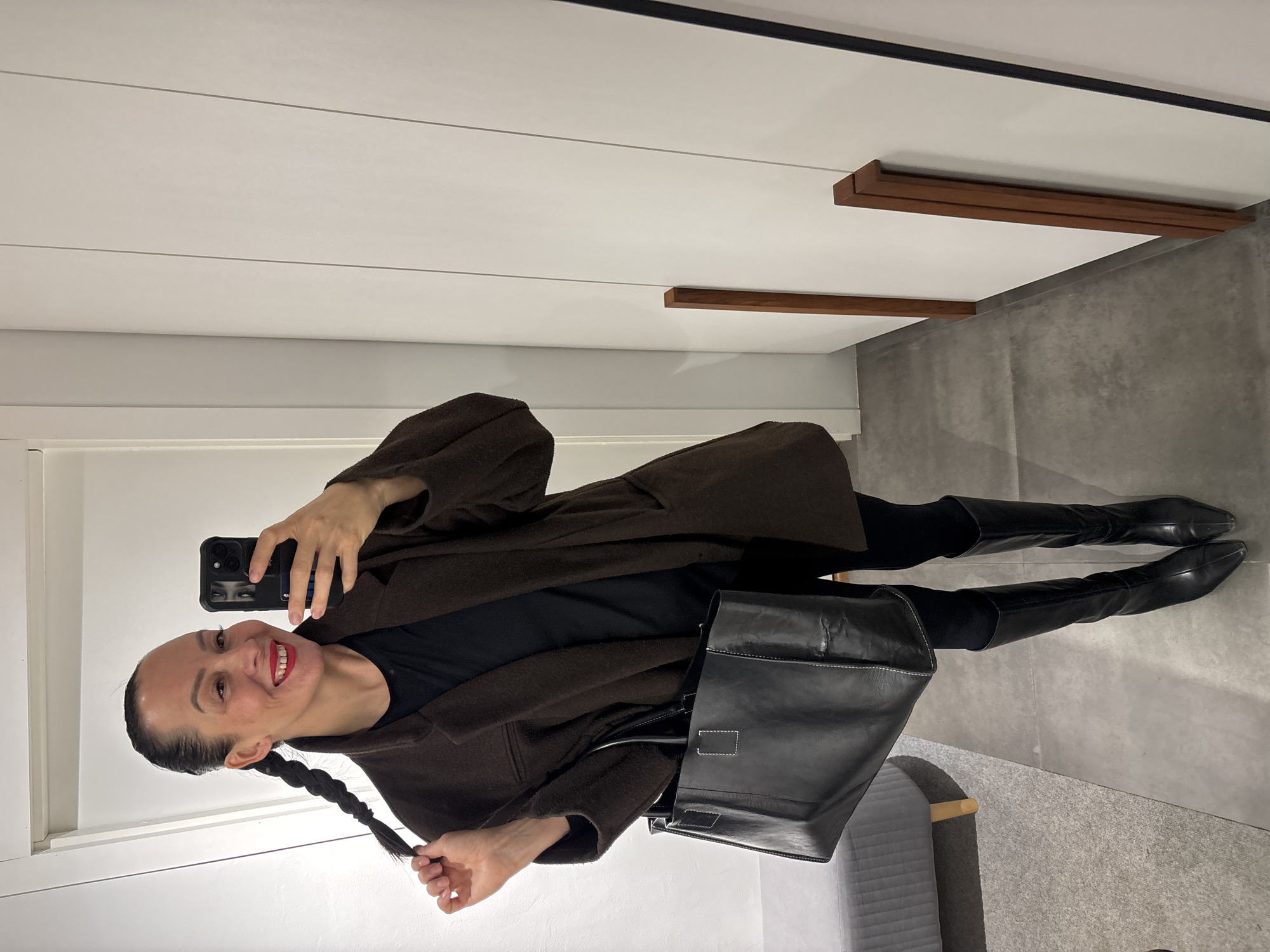 A woman is taking a selfie in a bathroom. She is wearing a brown coat, black top, black leggings, and black knee-high boots. She has a long braid, and is holding the end of it in her right hand. She is also carrying a black leather tote bag. The bathroom has white walls, grey tiled floors, and wooden trim. The location may be Chaikovsky, Russia.