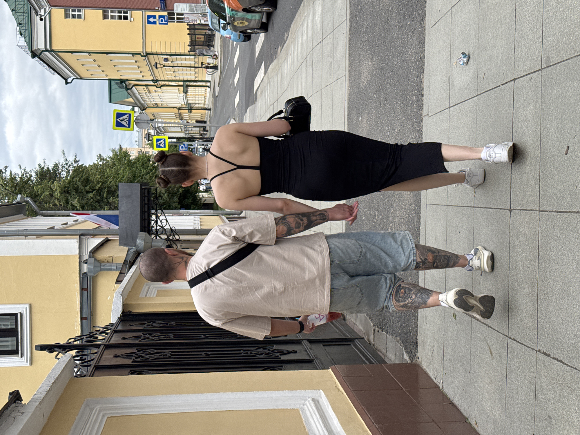 In Moscow, Russia, a man and a woman are walking hand-in-hand on a sidewalk next to a street with buildings and traffic. The man, from behind, wears a tan t-shirt, light blue denim shorts, and white sneakers. He has extensive tattoos on his legs and left arm, and is holding a clear plastic cup with a pink beverage. The woman, also viewed from behind, is wearing a black, form-fitting midi dress with spaghetti straps and white sneakers. She has her hair styled in two buns and is carrying a black handbag. The sidewalk is paved with gray concrete tiles. Behind them, a yellow building with a green roof is visible, along with a black ornate gate. Traffic signs, including pedestrian crossing signs, are present on the street. The sky is overcast.