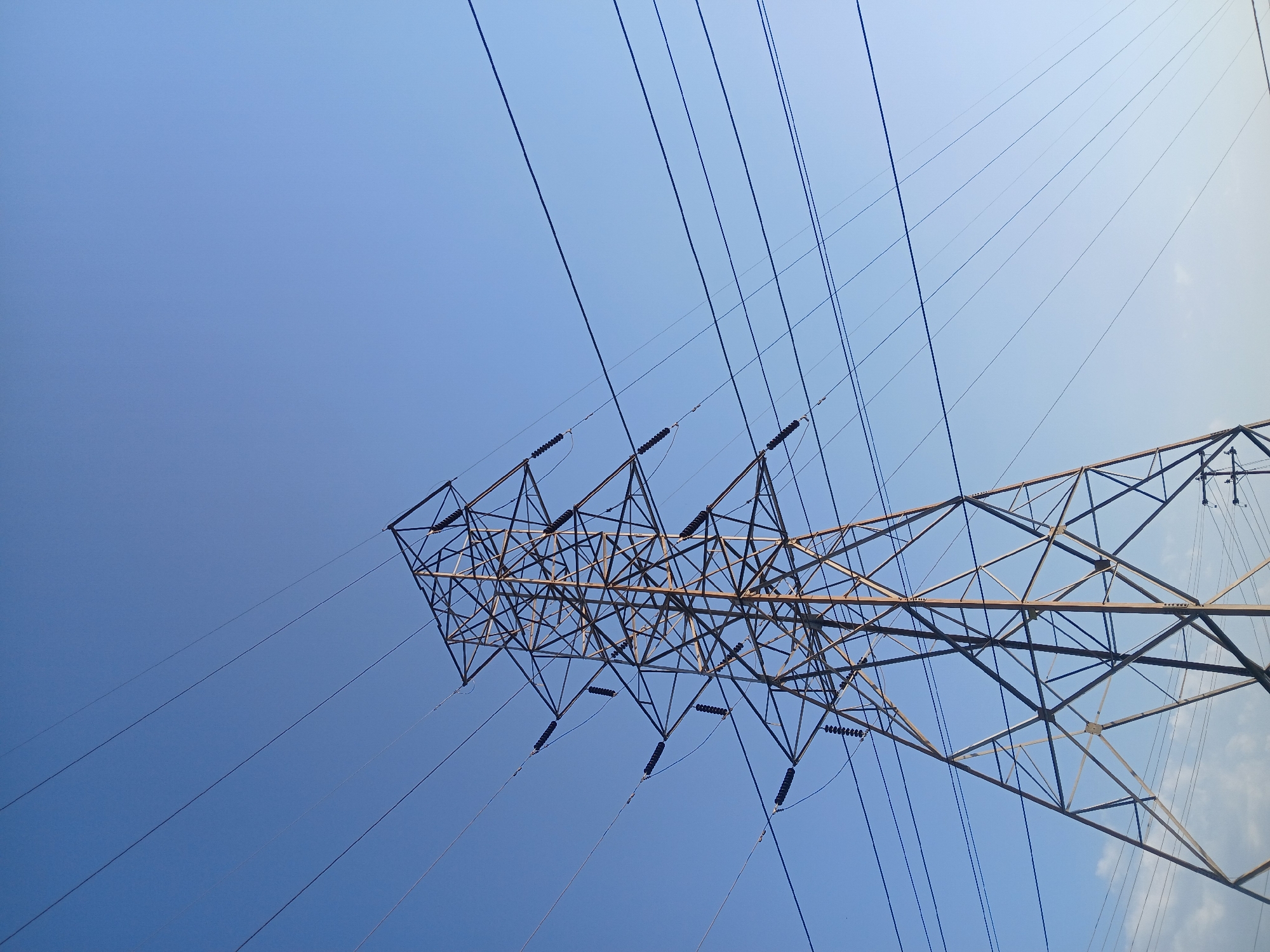 The image shows a low-angle view of a high-voltage electricity pylon against a bright blue sky. The pylon is constructed from a lattice of steel beams. Multiple power lines extend from the pylon, spanning across the upper portion of the frame. The image was taken in Ciudad Guayana, Venezuela.
