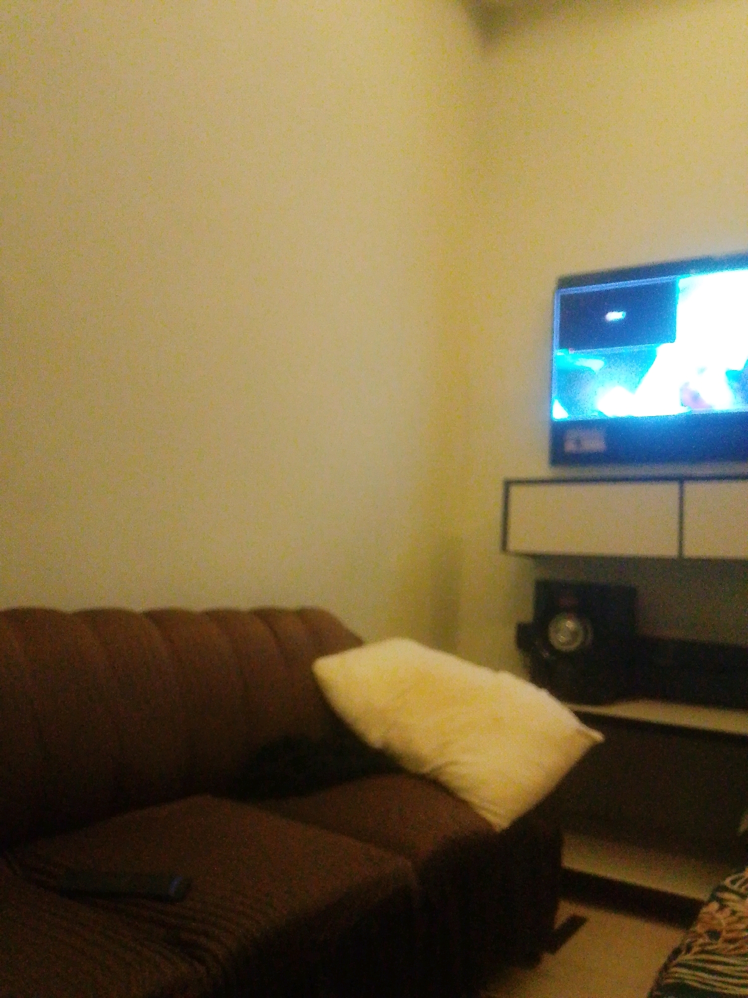 The image depicts a dimly lit living room scene, possibly in Faisalabad City Tehsil, Pakistan. A brown sectional sofa occupies the foreground, with a light-colored pillow resting on its right side. A remote control is visible on the seat of the sofa. In the background, a television is mounted on the wall above a dark entertainment unit. A black speaker is placed on the lower shelf of the entertainment unit. The walls are painted a pale yellow color. The overall atmosphere suggests a quiet, indoor setting.