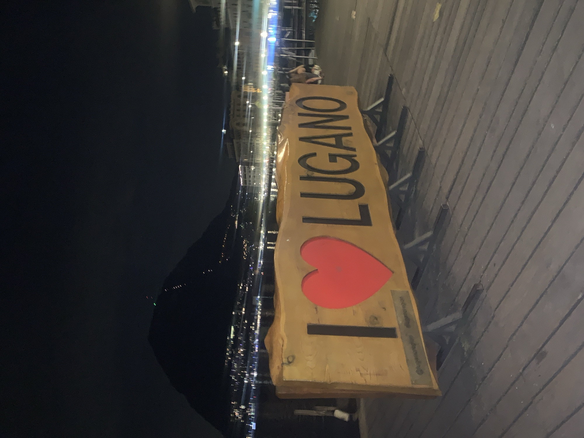 At night, a large wooden sign that reads "I LOVE LUGANO" is displayed prominently on a wooden boardwalk next to a body of water. The sign features a cutout red heart where the word "LOVE" would typically be. In the background, the city lights of Lugano, Switzerland, are visible, along with a dark mountain silhouette. The boardwalk is made of dark wooden planks.