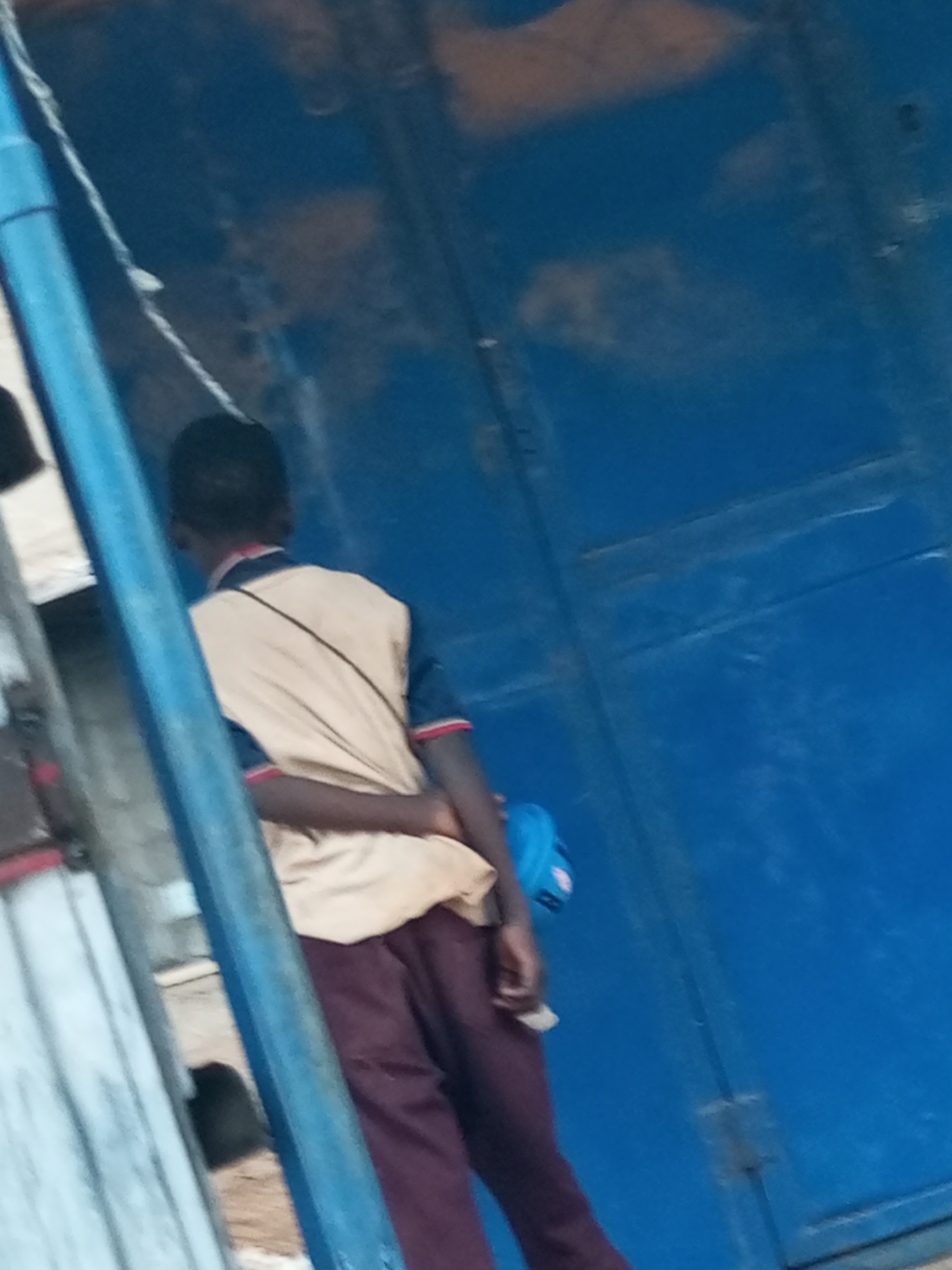 A person, viewed from behind and from the right, stands in front of a blue metal door in Tempure, Nigeria. The person wears a tan shirt with dark blue trim and maroon pants. A black strap is visible across their back, and a blue bag is attached to their right side. Their arms are crossed behind their back. The background consists of a textured blue surface, likely a wall or door, and a vertical blue pole or beam on the left side of the frame. The lighting suggests an outdoor environment during the day.
