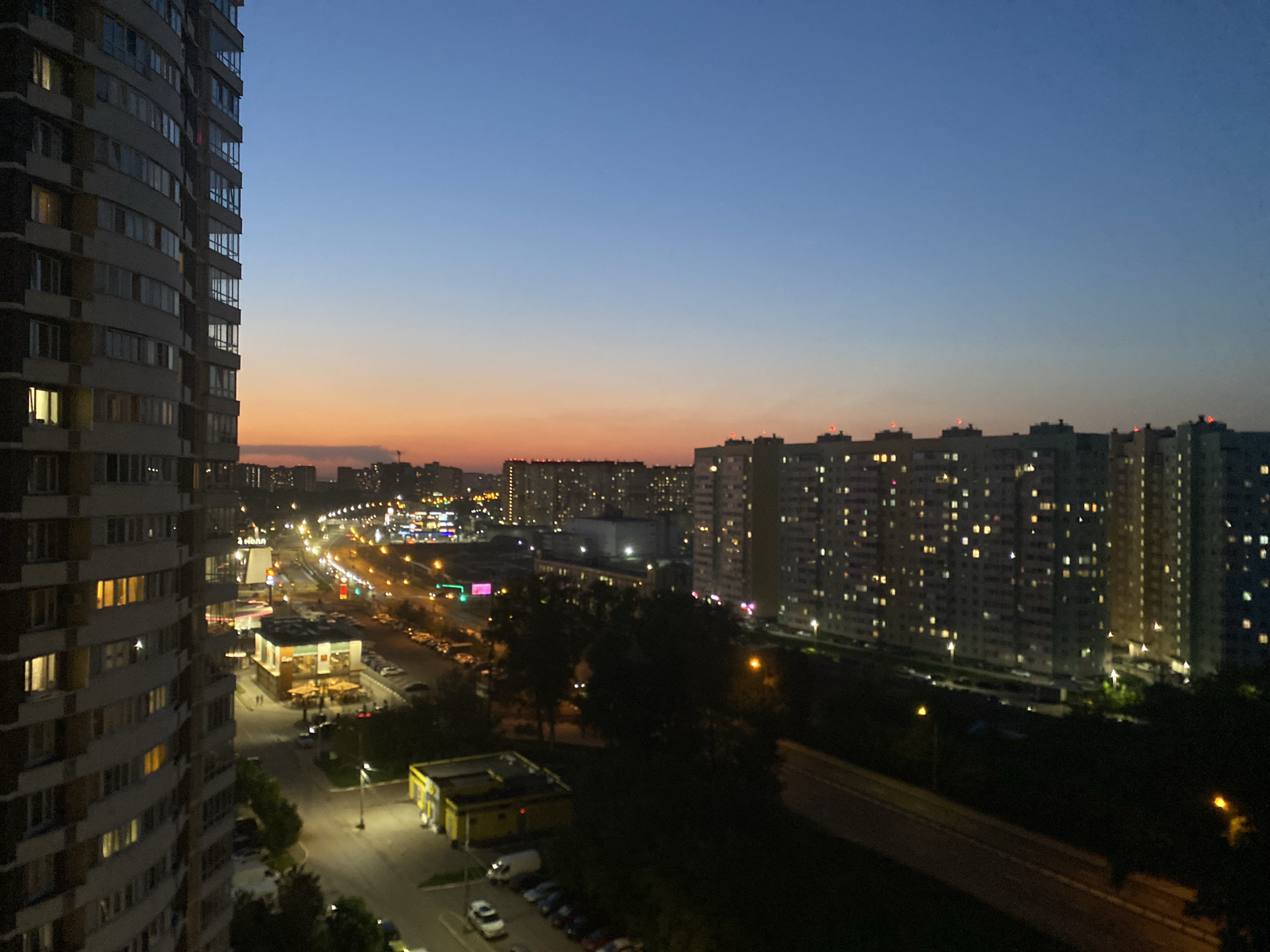 The image depicts an urban landscape at dusk, likely in an area with high-density residential buildings. A tall apartment building dominates the left side of the frame, with numerous windows, some illuminated, suggesting occupancy. In the distance, a cluster of similar high-rise buildings extends across the horizon, also with many illuminated windows.
Below, a streetscape includes a brightly lit commercial area with storefronts and parked vehicles. Streetlights illuminate the roads and sidewalks. Trees and vegetation are visible, interspersed among the buildings and along the roadways.
The sky transitions from a deep blue at the top to a soft orange near the horizon, indicating twilight. The overall environment suggests a densely populated residential area with active commercial zones. No people are clearly visible.