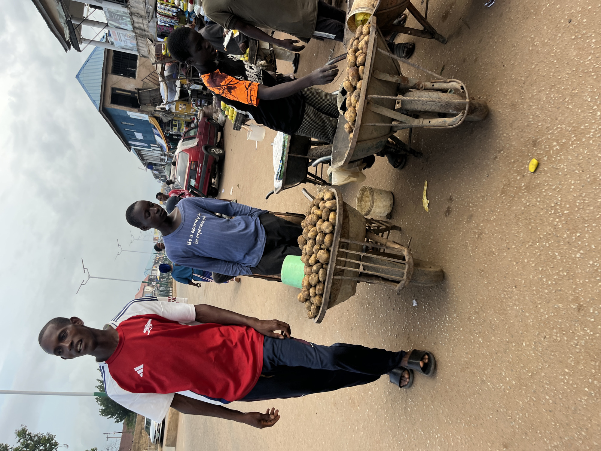 In Bukuru, Nigeria, a street scene unfolds with several individuals engaged in commerce. A man wearing a red and white athletic shirt and dark pants walks on the left side of the frame, looking to his left. Next to him, a wheelbarrow filled with potatoes sits on the sandy ground. Behind him, another young man in a blue t-shirt that reads "Life is a journey to be experienced" is visible. In the foreground, a second wheelbarrow, also laden with potatoes, is positioned in front of a street vendor wearing a black t-shirt with orange accents. This vendor is actively engaged with the potatoes. Another wheelbarrow further back contains citrus fruits. The environment is characteristic of a bustling market with buildings lining the street, some with corrugated metal roofs, and various goods displayed for sale. The sky is overcast.