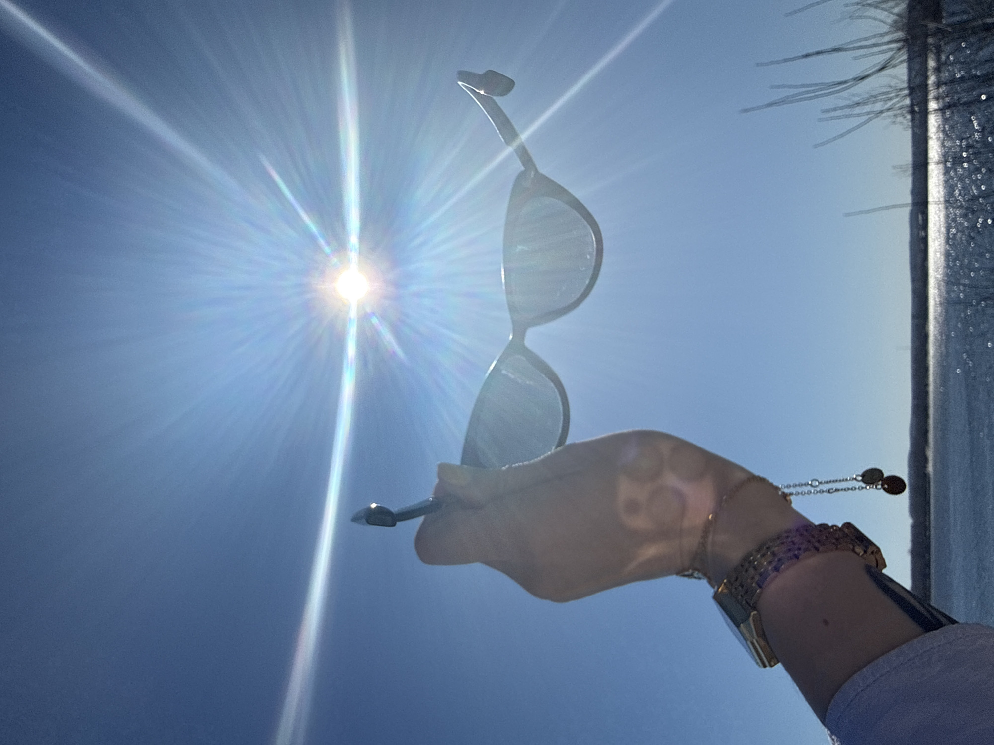 A photograph taken outdoors in Chaikovsky, Russia shows a person holding up a pair of sunglasses toward a bright sun in a clear blue sky. The sun is positioned in the upper left of the frame, radiating light. The sunglasses are held in the person's hand, which is adorned with a bracelet, a watch, and what appears to be a small tattoo or design. To the right of the person's arm is a vertical structure that is partially visible.