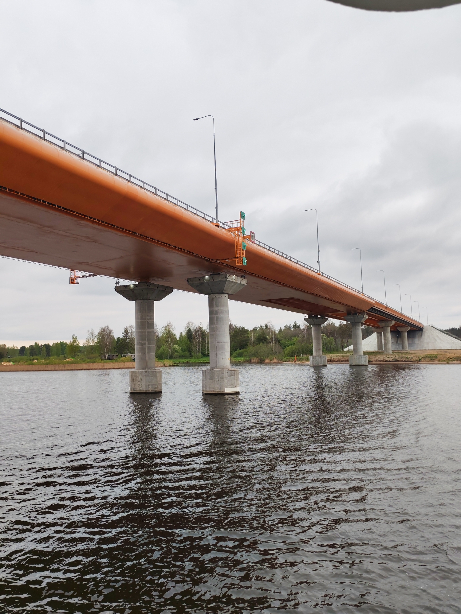 The image depicts a modern orange-colored bridge spanning a body of water, likely a river. The bridge has concrete support pillars resting in the water. There are lampposts lining the bridge's roadway. The sky is overcast. Vegetation is visible on the riverbank in the background. The water surface is rippled. The location is the city of Orsha, Russia. No people are visible.