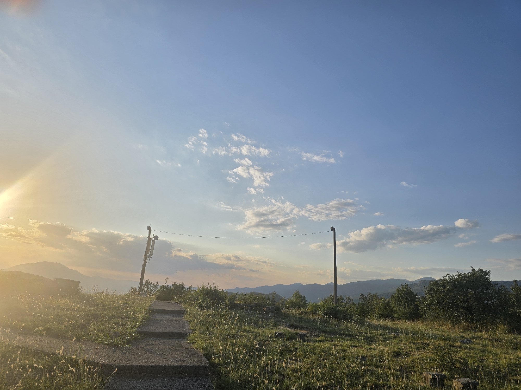 The image depicts an outdoor scene at sunset, showing a concrete pathway leading upwards into a grassy, hilly area. The pathway is flanked by tall, dry grass and some scattered bushes. In the background, a range of mountains is visible under a vast blue sky with scattered white clouds. To the left, the setting sun casts a warm, golden light and lens flare across the scene, illuminating the landscape and creating a hazy atmosphere. A single utility pole with what appears to be communication equipment is situated along the pathway, and another lamppost stands further along the path, towards the right side of the image. There are no people visible in the scene. The environment suggests a rural or elevated natural setting, possibly a hiking trail or viewpoint.
