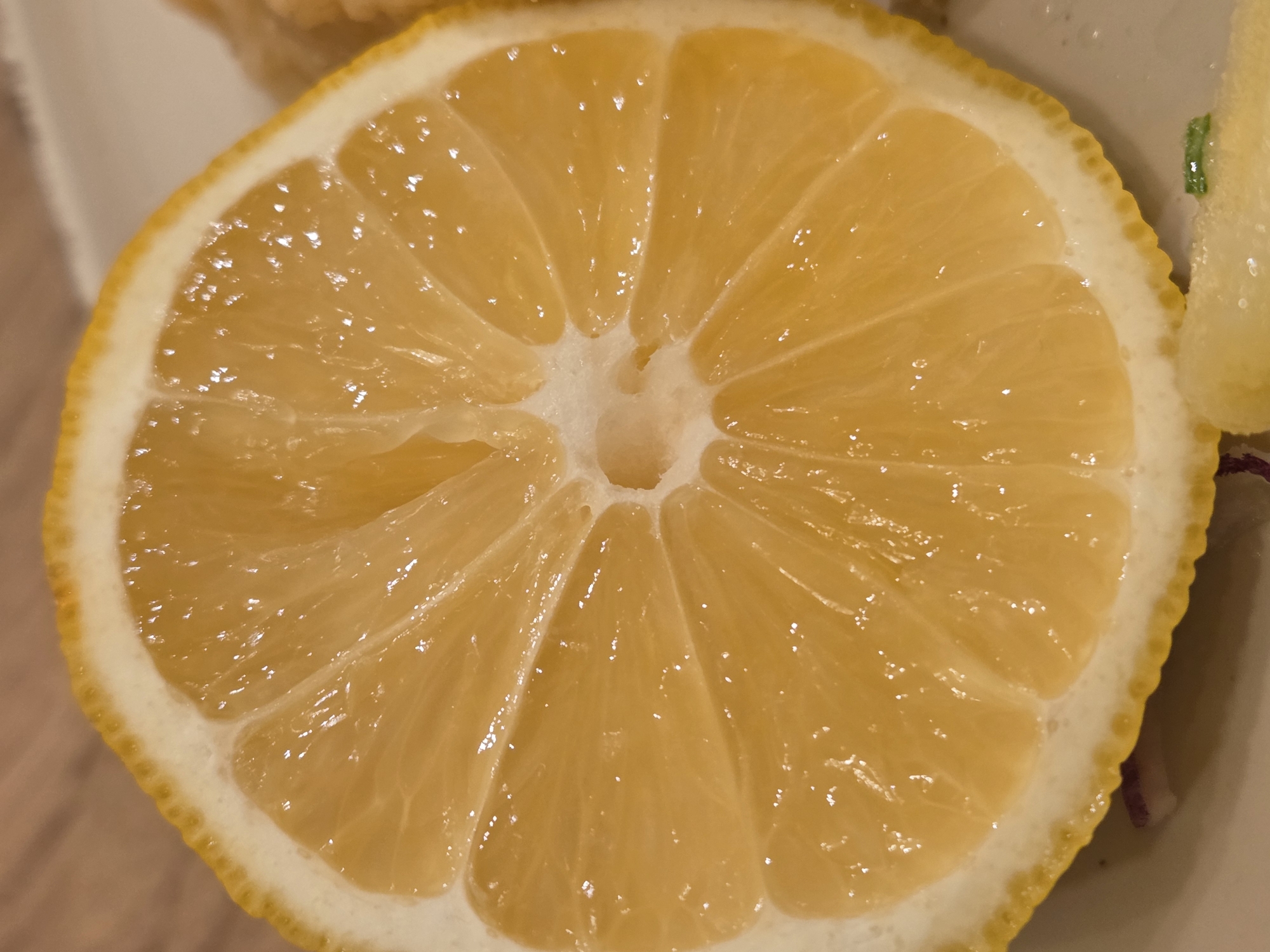 A close-up photograph shows a cross-section of a lemon. The lemon is cut in half and displays its segments radiating from a central white core. The segments are a translucent yellow, with visible juice. The rind of the lemon is thick, with a white pith and a thin yellow outer layer. The lemon appears to be sitting on a plate, with some other food items partially visible in the periphery. The context of the location in Varna, Bulgaria, is not discernible from the image.
