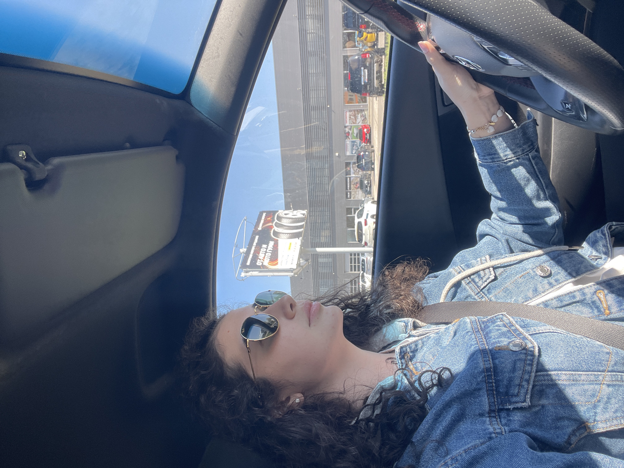 A woman with curly brown hair and sunglasses is in the driver's seat of a car, wearing a denim jacket and a seatbelt. Her right hand is visible on the steering wheel. The car has a sunroof. Outside the car window, there is a building with multiple floors and parked cars. A billboard is also visible. The location is Sofia, Bulgaria.