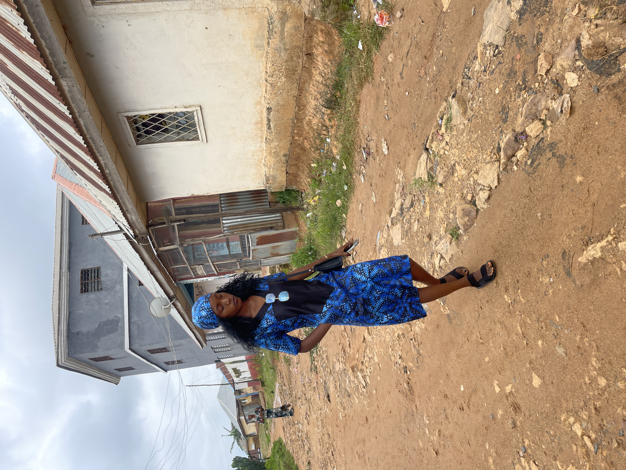 A young woman stands on a dirt path in front of a building, her body angled slightly away from the camera. She is wearing a blue and black patterned dress with a matching headscarf. She has long, dark, curly hair and is wearing sunglasses perched on her head. She is holding a black bag in her left hand. The background shows a building with a corrugated metal roof and several windows. The ground is a mixture of dirt, rocks, and sparse green grass. The sky is overcast. The scene suggests an outdoor setting, possibly in a residential or commercial area. The lighting indicates it is daytime.