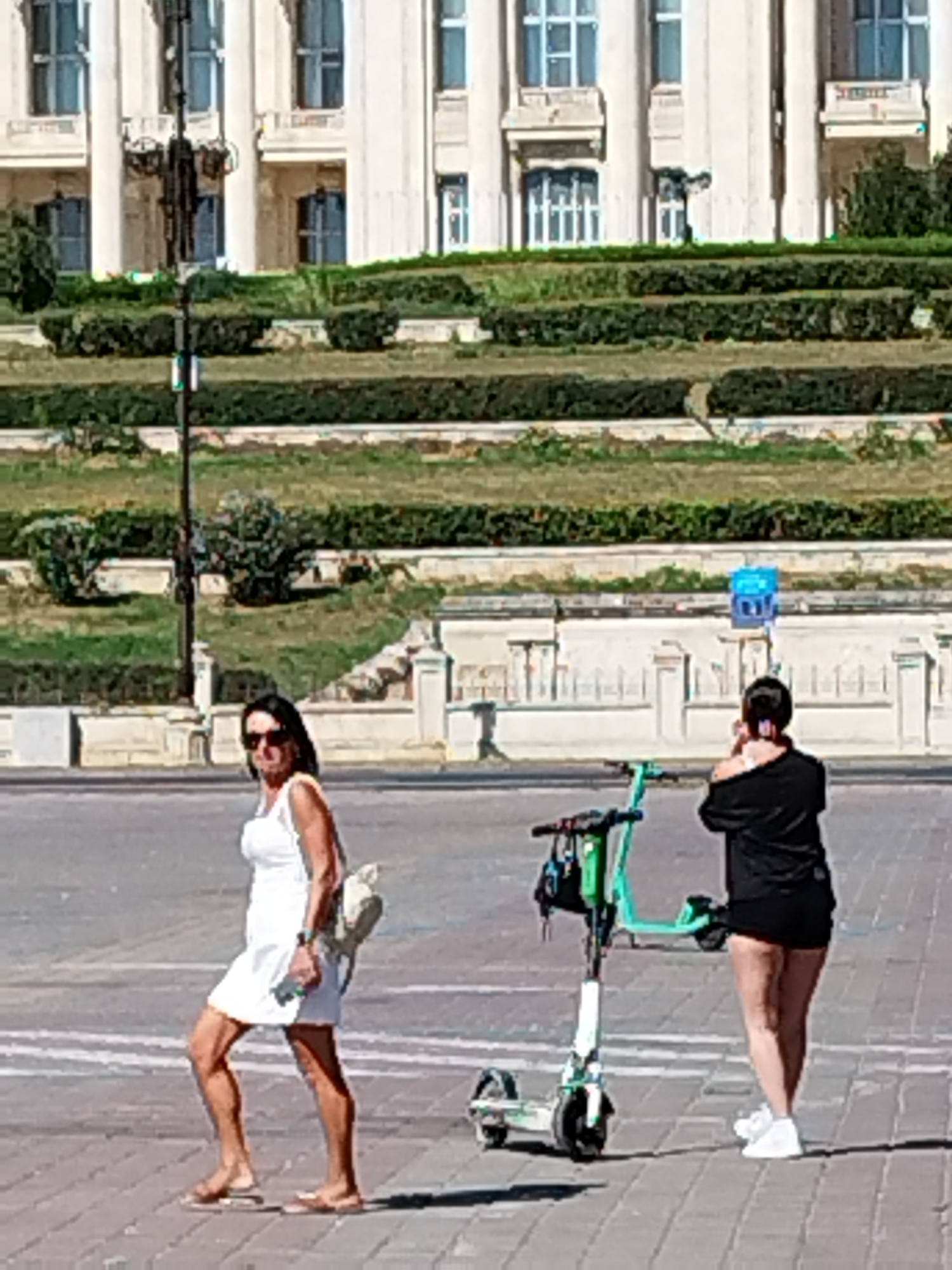 Two women are walking on a paved area in front of a large building with a manicured lawn and trees. The building appears to be a governmental or historical structure. On the right, a woman in a black t-shirt and shorts is holding her phone to her ear. In the foreground, to the left, a woman in a white dress and sandals is walking away from the camera, carrying a light-colored bag over her shoulder. A green electric scooter is parked to the right of the woman in black. The scene is consistent with an outdoor public space in Bucharest, Romania, possibly near the Palace of the Parliament.