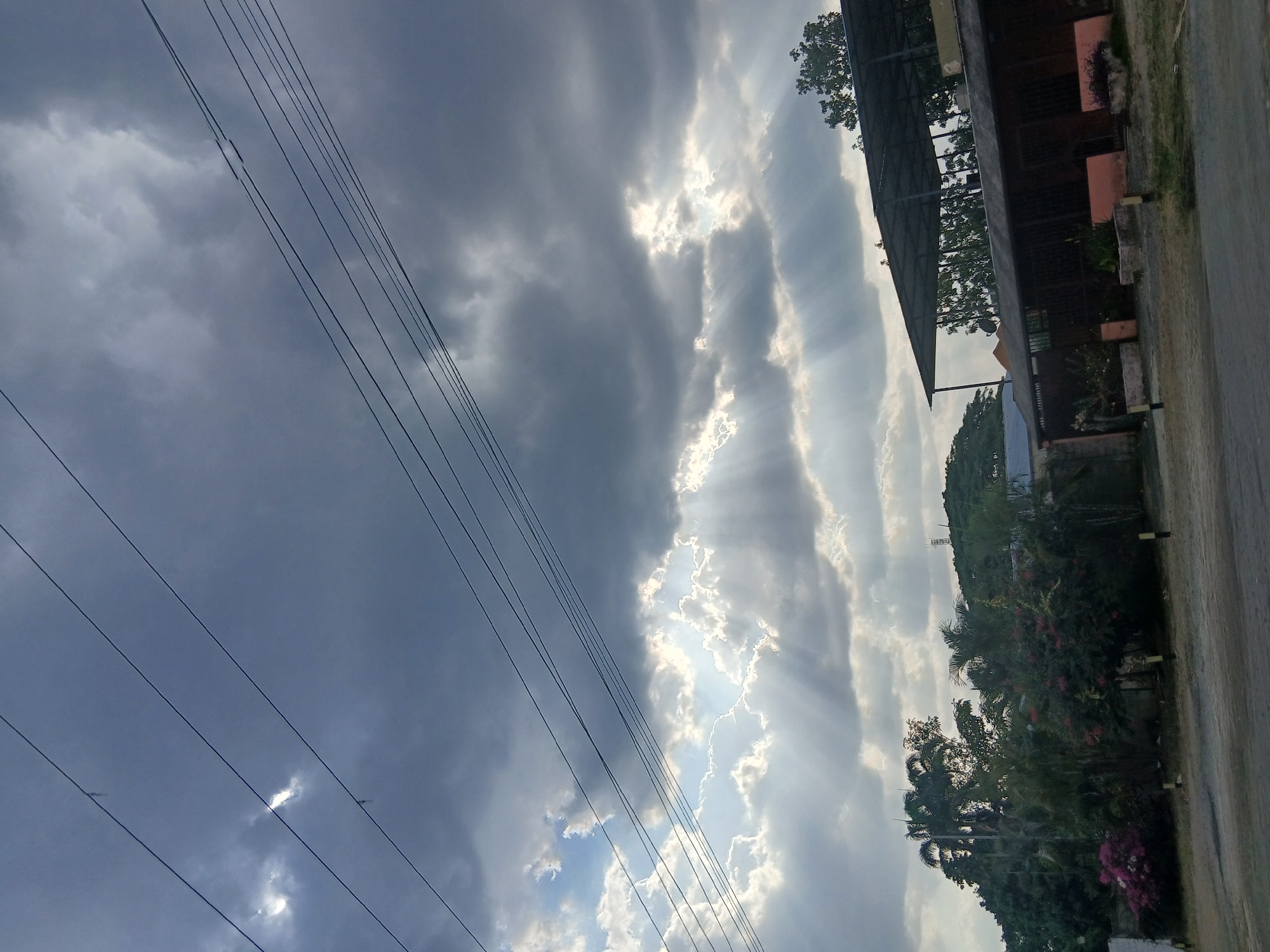 The image shows a view of the sky with scattered clouds, some dark and ominous, others allowing sunlight to break through in rays. Electrical power lines stretch across the sky. On the right, there's a glimpse of buildings, vegetation, and a road. Based on the context clues of the user prompt, it can be inferred that this scene is in Tinaquillo, Venezuela. No people are visible.