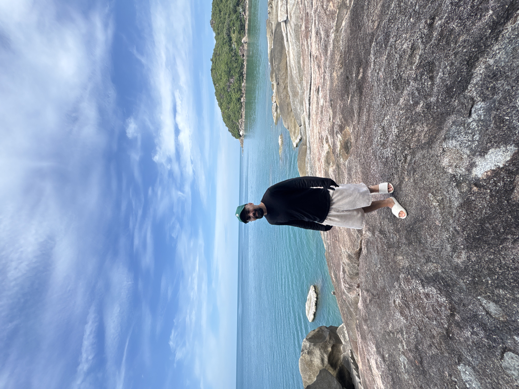 A man stands on a rocky outcrop overlooking the ocean. He is wearing a black long-sleeved shirt, light-colored shorts, a green baseball cap, and white slides. He is facing the camera with a slight smile. The ocean is a vibrant turquoise color, and the sky is a bright blue with scattered white clouds. In the background, a lush green island juts out of the water. The rocks in the foreground are large and uneven, with some patches of moss and lichen. The scene appears to be a coastal area in Ko Samui District, Thailand.