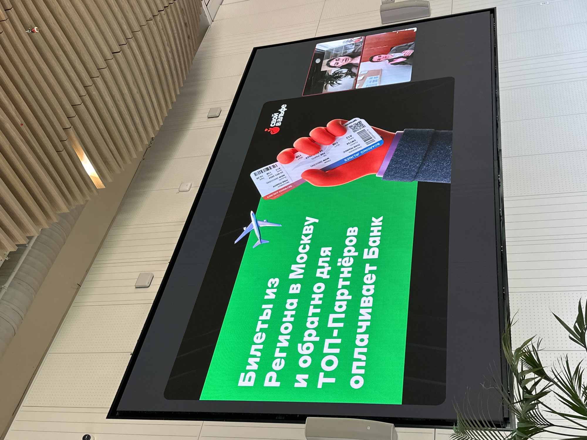The image shows a large digital display mounted on a wall, likely indoors, possibly in a public space. The display shows an advertisement, with text in Russian: "Билеты из региона в Москву и обратно для ТОП-Партнёров оплачивает Банк" ("Tickets from the region to Moscow and back for TOP-Partners paid by the Bank"). The advertisement also shows a hand holding airline tickets. There is a video call displayed in the upper section of the display. To the left of the display is a wooden slat wall. A plant is located to the right of the display. Given that the advertisement is in Russian, it is most likely located in Russia, and the text is advertising something related to tickets to Moscow, suggesting a location in Moscow.