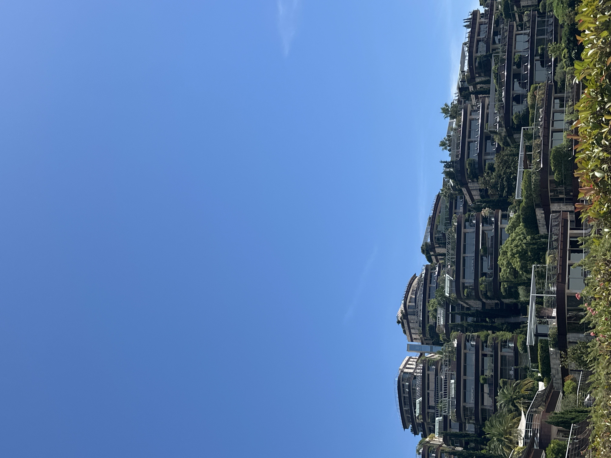 The image shows a cluster of modern, multi-story buildings with curved balconies and extensive greenery cascading down their sides, designed to blend with the natural landscape. The architecture features large windows and dark-colored accents. The buildings are situated on a hillside, suggesting a coastal or elevated location. The sky above is a clear, vibrant blue with a few wispy clouds visible on the right side. In the foreground, lush green and red foliage is prominent, hinting at a well-maintained garden or park area. The overall scene depicts a residential or hotel complex with an emphasis on contemporary design and integration with nature. This is the complex "Dukley Gardens" located in Budva, Montenegro.
