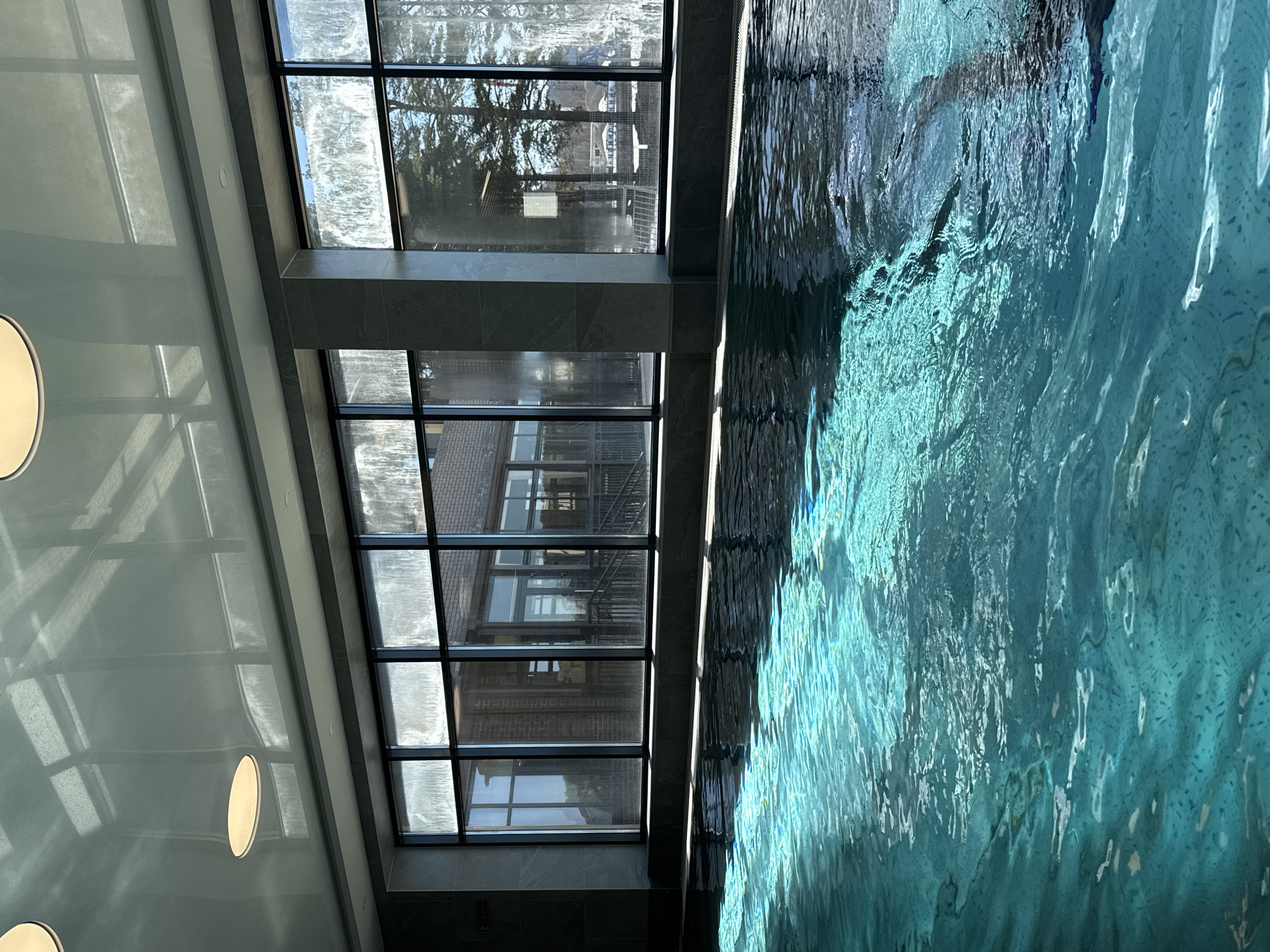 The image depicts an indoor swimming pool, with a view through a multi-pane window. The water in the pool is a vibrant blue, with ripples and surface reflections creating patterns of light and dark. Outside the window, a building is visible, including a brick wall and metal staircase. Reflected in the window are trees and a glimpse of the sky. The architecture of the pool area features a light-colored wall with round light fixtures. The scene does not contain any identifiable people or indications of city location.