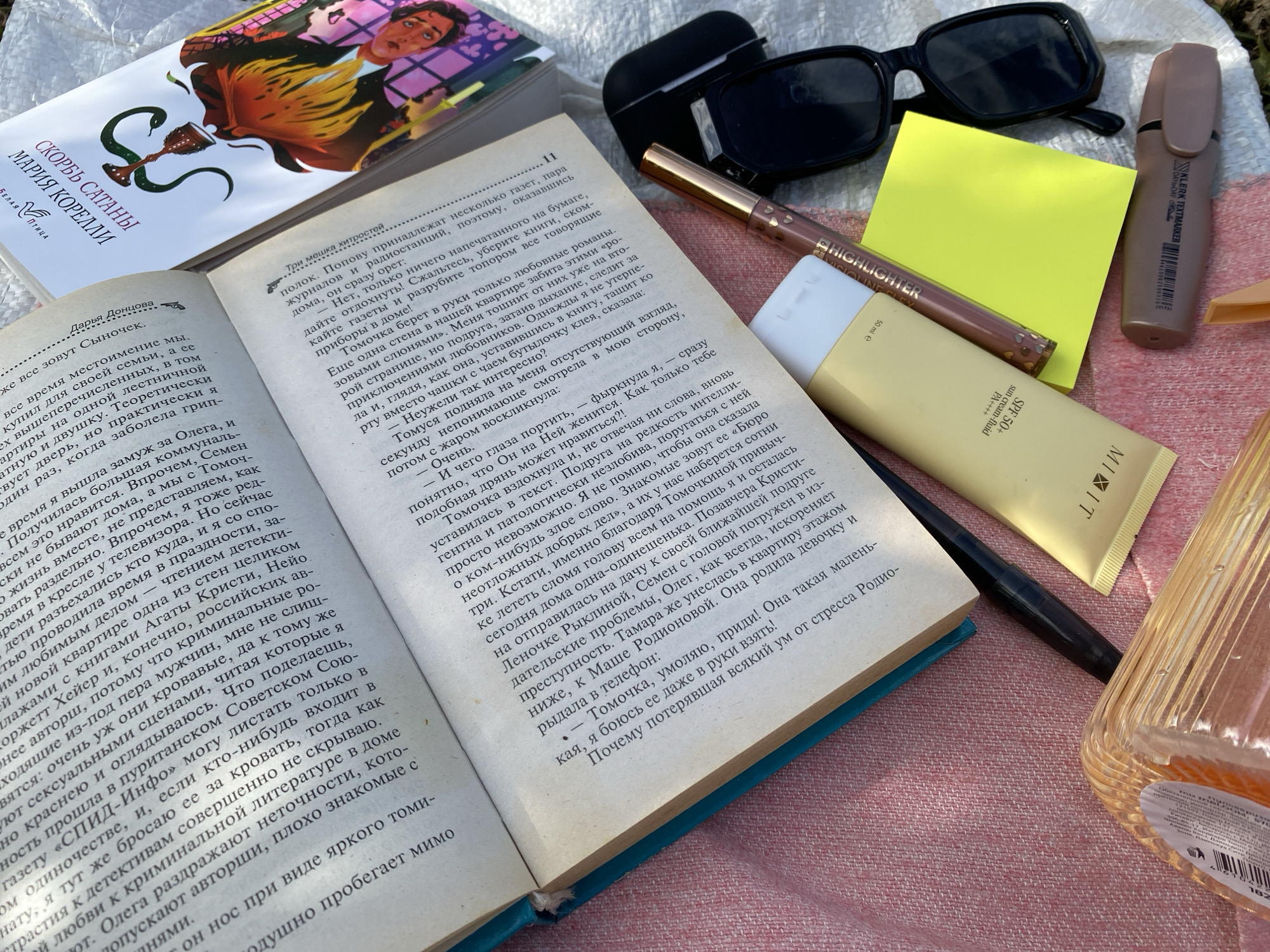 The image shows an open book with Russian text on a textured pink surface, possibly a blanket or fabric. The book is positioned at an angle, with the title "СКОРБЬ САТАНЫ" and author "МАРИЯ КОРЕЛЛИ" visible on the cover of a separate book laying on top of the open pages. Several other items are scattered around the book: a pair of black sunglasses, a highlighter pen, a tube of sunscreen labeled "SPF 50+", and a brown marker or pen. The scene suggests a relaxed outdoor setting, possibly a picnic or a day spent reading outside. The location is identified as Elovo, Russia.