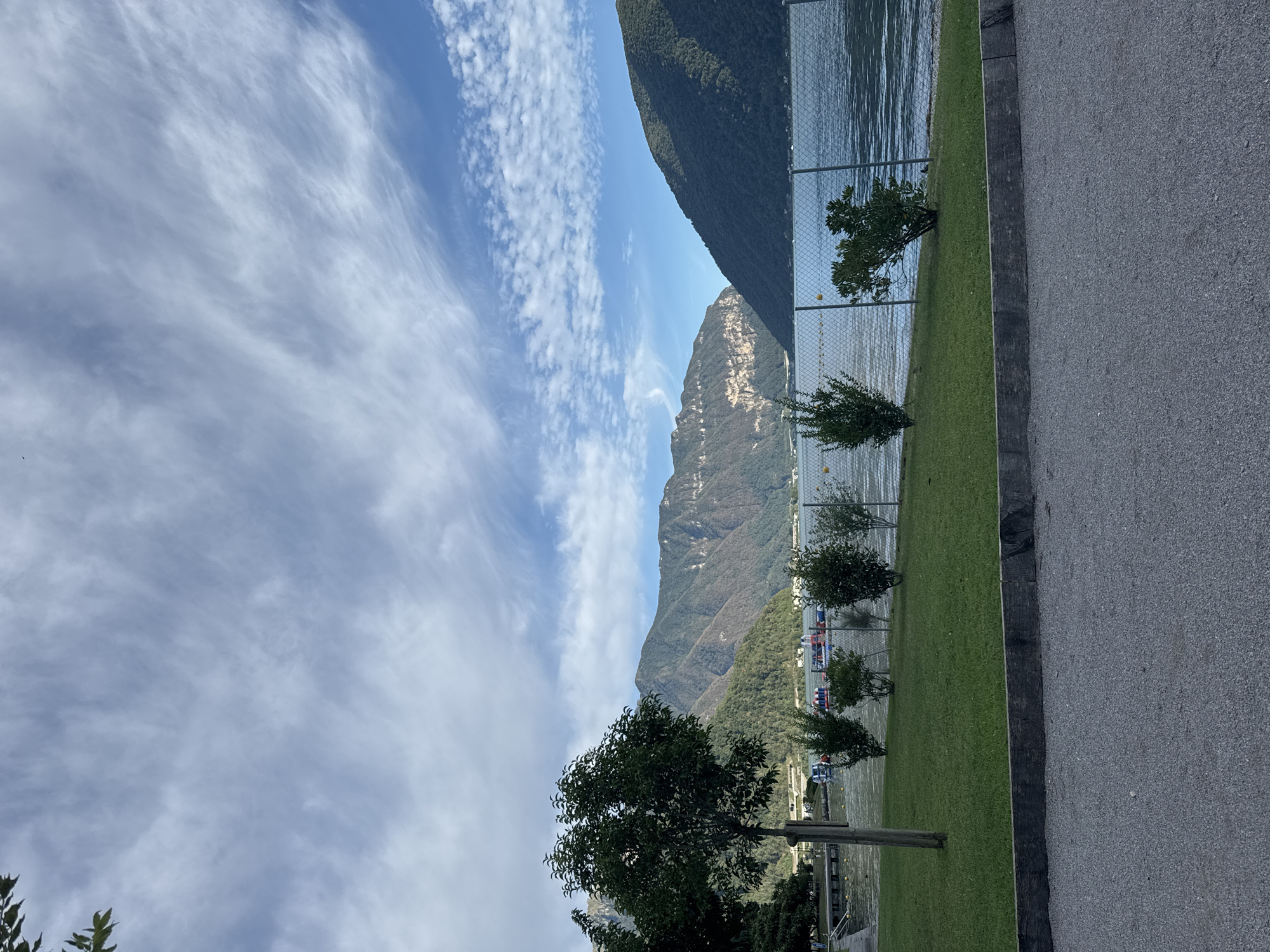 A vertical, eye-level, medium shot depicts the lakeside in Melide, Switzerland, on a clear day with a partly cloudy sky. In the foreground, a dark grey asphalt path runs horizontally across the bottom of the frame, separated from a neat strip of vibrant green grass by a weathered wooden beam. Beyond the grass, a chain-link fence runs parallel to the shore, with a few small, leafy green bushes scattered along its base.
The middle ground is dominated by the expansive, rippling blue water of Lake Lugano. On the far side of the lake, a range of majestic mountains rises steeply. The slopes of these mountains are densely covered in a patchwork of green trees, with some areas showing lighter, rocky surfaces. The sky above is a brilliant blue, streaked with wispy white clouds that are illuminated by the sun.
Along the edge of the lake, a wooden pier extends into the water, and several small boats, some with people visible, are anchored near the shore. In the distance, a few more boats and structures are visible along the shoreline. The overall atmosphere is serene and picturesque, showcasing the natural beauty of the Swiss Alps and the tranquil lakeside.