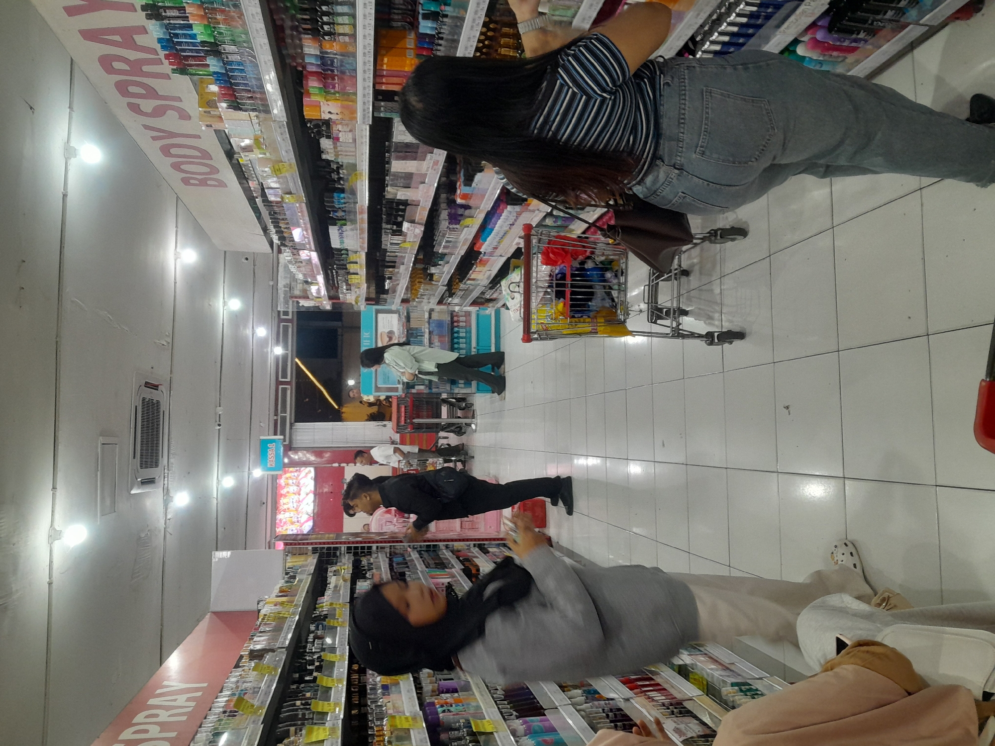 The image depicts the interior of a brightly lit supermarket aisle, likely in Banyuwangi, Indonesia, given the context provided. The aisle is lined with shelves stocked with numerous personal care products, primarily bottles of body spray and other cosmetics. The shelves are organized and well-maintained, with products neatly arranged.
Several individuals are present in the scene, shopping and browsing the merchandise. In the foreground, a woman wearing a grey top and black hijab is looking at the products on a shelf. To her right, a man in a black shirt and pants is also examining items. Further down the aisle, another woman in a light green shirt and dark pants is seen looking at products on a different shelf. In the mid-ground, a shopping cart is visible, laden with various items, including what appears to be a red bag. In the background, other shoppers and store employees can be seen, with some individuals near a checkout counter.
The flooring is tiled in a light color, and the ceiling features fluorescent lighting, contributing to the overall bright ambiance. Promotional signage is visible on the upper parts of the shelves, indicating product categories such as "BODY SPRAY". The environment suggests a typical retail setting where customers are engaged in shopping activities.