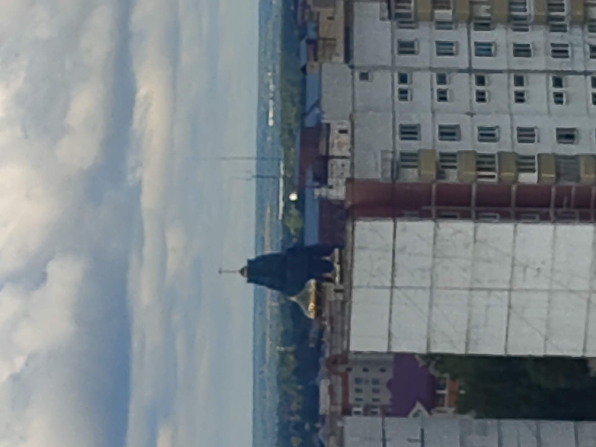 A wide shot, slightly blurry, shows a cityscape under a cloudy sky. Dominating the center of the frame is the dark green spire of a building, topped with a golden dome and a cross. To its left, another smaller golden dome is visible. In the foreground and mid-ground, rows of multi-story residential buildings with light-colored facades and visible windows line the cityscape. Some buildings have balconies, and one features a visible burgundy-colored roof. The background shows a hazy expanse of trees and more distant structures. The image captures an urban environment with a prominent religious building.