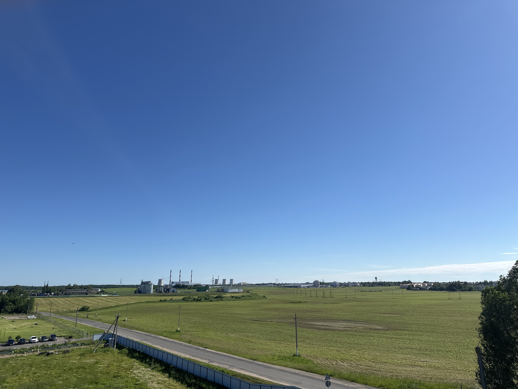 This is a wide shot of a rural landscape under a clear blue sky. The scene is dominated by a vast expanse of green fields, some of which appear to be cultivated with rows of young crops. A paved road runs diagonally across the lower half of the image, bordered by a light-colored fence. On the left side of the road, a parking lot is visible with several cars parked, and a white car is driving away. In the distance, a cluster of industrial buildings with tall smokestacks and cooling towers stands against the horizon, suggesting a power plant or factory. Further to the right, a small town with residential houses can be seen nestled amongst trees. Several wooden poles and power lines are scattered throughout the fields, indicating the presence of infrastructure. The time of day appears to be daytime, likely morning or afternoon, given the bright sunlight casting minimal shadows. The weather is clear and sunny, with no clouds visible in the sky. The overall impression is one of a peaceful, somewhat industrialized rural setting in Belarus.