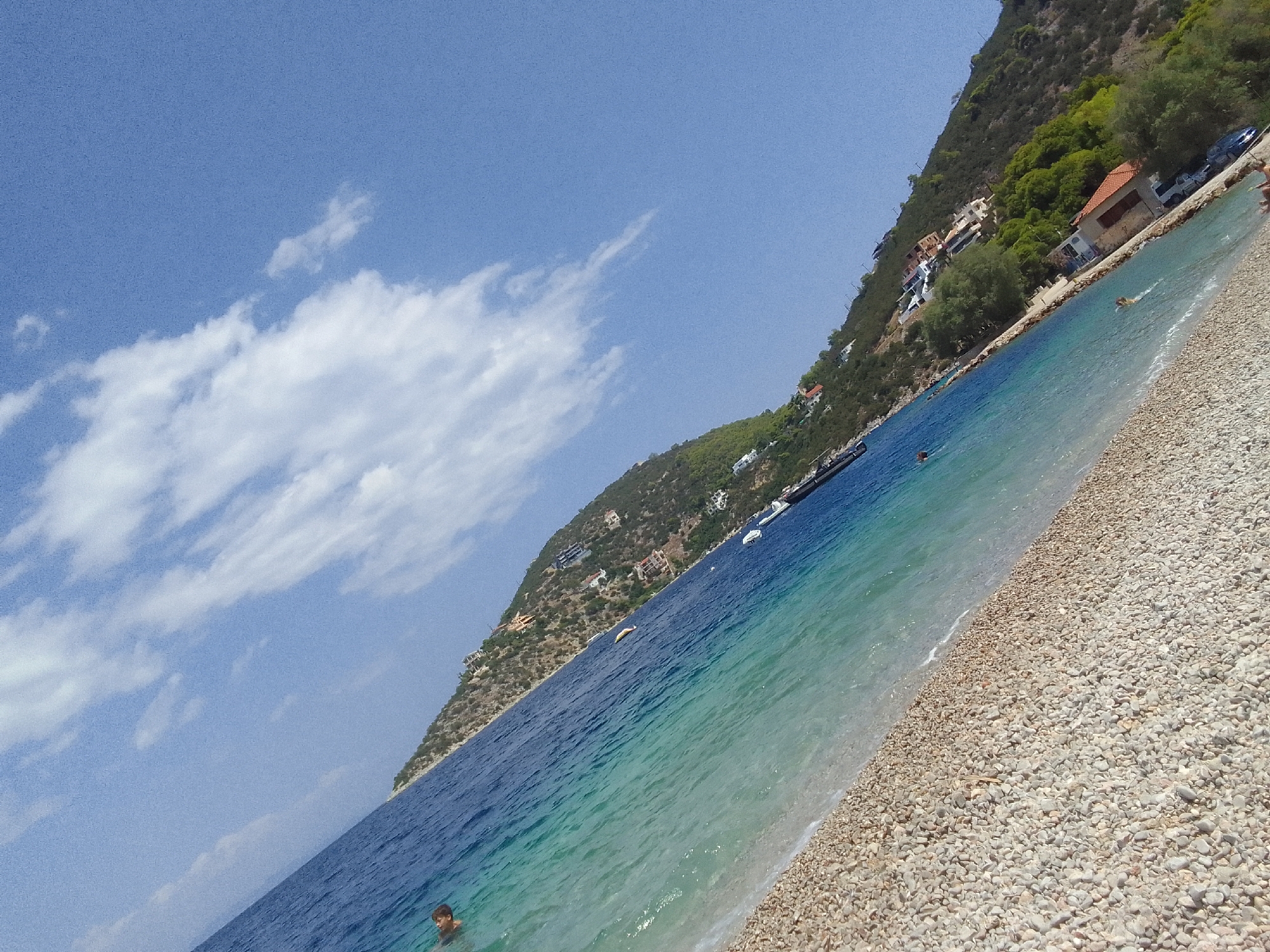 The image shows a coastal scene in the Municipal Unit of Solygeia, Greece. A steep, tree-covered hillside slopes down to a pebble beach and the clear blue-green waters of the sea. Several houses are visible nestled into the hillside. The sea is calm, with a few boats anchored offshore. A person is swimming in the water. The sky is blue with scattered white clouds. The perspective is from a low angle, looking up at the hillside.