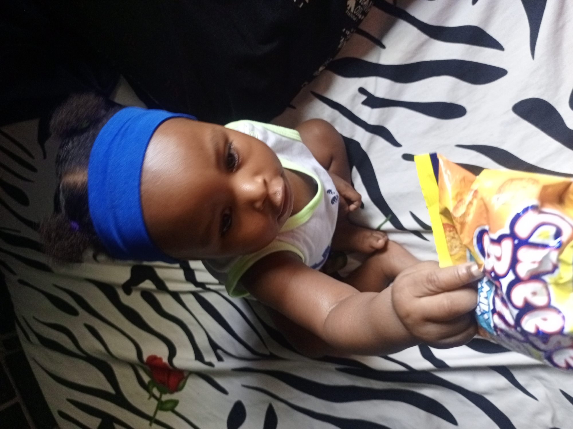 This image captures a baby, likely in an indoor setting in Keffi, Nigeria. The baby is positioned on a surface covered with a white and black zebra-striped sheet, which also displays a red rose design in the bottom left corner. The baby has dark skin and is wearing a bright blue wide headband and a white sleeveless top with lime green trim. Their dark hair is partially visible beneath the headband. The baby looks up towards the camera or an unseen individual, their expression appearing focused and curious. The baby's right hand is grasping a yellow and orange snack bag, on which the text "Swee" is prominently visible in blue and white letters, suggesting a sweet treat. The lighting suggests it is daytime.