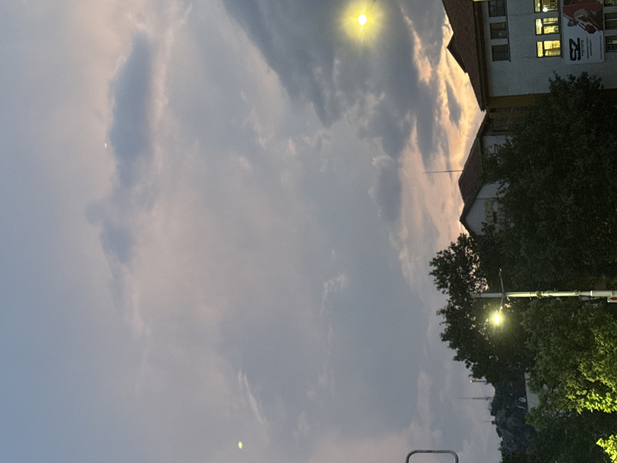 The image depicts a view of Plovdiv, Bulgaria, with a dusky sky filled with clouds. In the foreground, a streetlamp illuminates the scene. Behind the lamp, lush green trees obscure much of the view of buildings and a rocky hill in the distance. The hill features several communication towers. To the right of the image, the upper floors of two buildings are visible. The building on the right is white with a tiled roof and windows that are lit from within. A large sign with the letters "SZ" is visible on this building. The building on the left is also partially visible, with some graffiti on its facade. The overall impression is of a twilight scene in an urban setting, with a blend of natural elements and man-made structures.