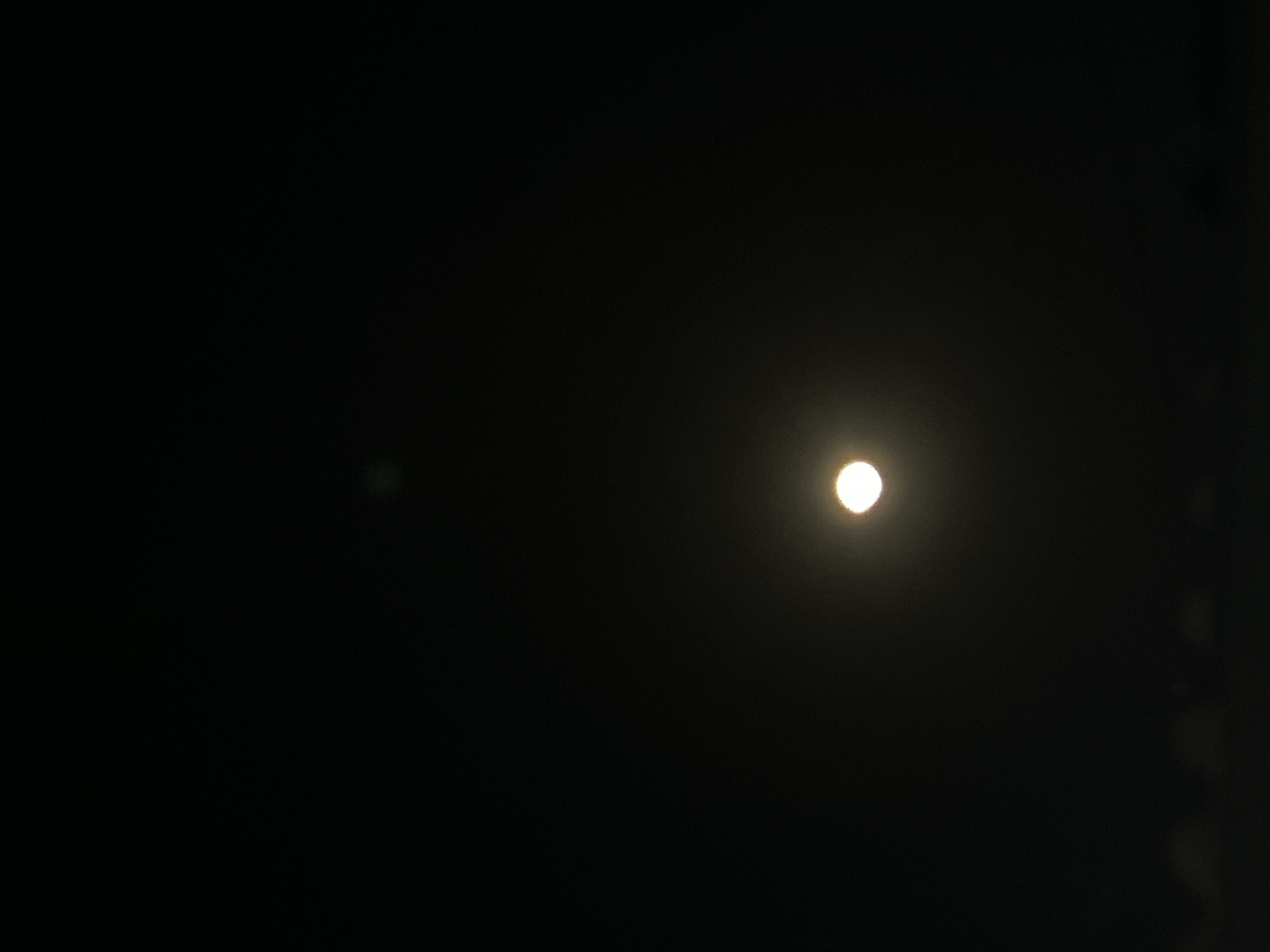 The image depicts a dark night sky with a bright, circular moon located in the lower center. The moon is luminous and appears to be slightly hazy, suggesting the presence of clouds or atmospheric conditions. Below the moon, a faint, dark silhouette of what could be the horizon or distant structures is visible. In the upper portion of the sky, a very faint, small, diffused light source is present. The overall scene is dominated by darkness, with the moon being the primary source of illumination. There are no visible people, animals, or specific man-made objects other than the potential distant silhouette. The location in Denpasar, Indonesia, is not visually identifiable from the provided image.
