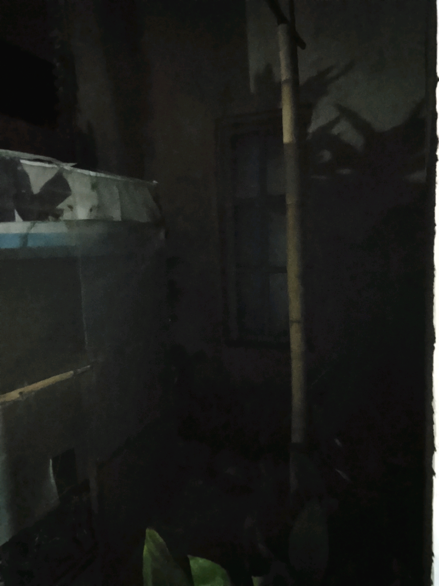 The image depicts a dark, indoor environment, possibly a dilapidated room or shed, with a focus on shadowed areas. A prominent vertical bamboo pole stands near the center, casting distinct shadows on the textured wall behind it. These shadows suggest the presence of objects or possibly hands, forming shapes that resemble animal ears or horns. To the left, a structure with a blue and white striped surface is partially visible, suggesting some sort of shelf or enclosure. A small, yellow pole or stick extends horizontally from the lower left towards the center. The overall impression is one of neglect and low lighting, with the shadows creating an ominous or mysterious atmosphere. The location is identified as Ujungrusi, Indonesia.