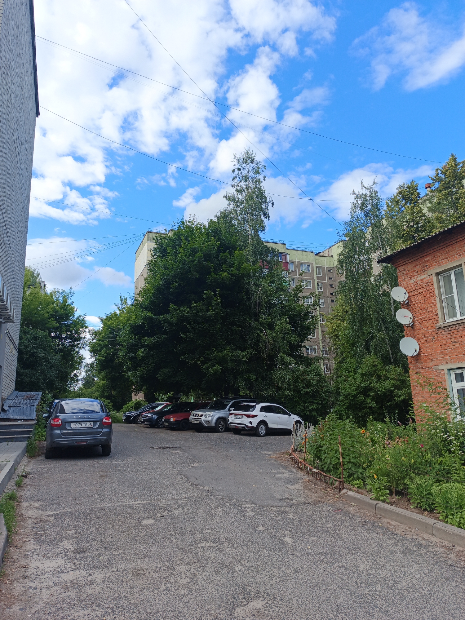 A sunny day in Kursk, Russia, as evidenced by the blue sky dotted with white, fluffy clouds. The scene is an urban setting, likely a residential area, featuring a paved courtyard with several cars parked. A dark gray Lada sedan is prominently parked in the foreground, facing away from the viewer, with its license plate clearly visible. Behind it, a line of other vehicles, including a white SUV, are parked. The courtyard is bordered by a lush green area with trees and flowering plants, suggesting a well-maintained outdoor space. To the right, a brick building with white-framed windows stands, adorned with three satellite dishes mounted on its facade. A multi-story apartment building is visible in the background, partially obscured by trees. Power lines crisscross the sky, a common sight in urban landscapes. The overall impression is one of a typical day in a Russian city, with a focus on the everyday life of its residents.