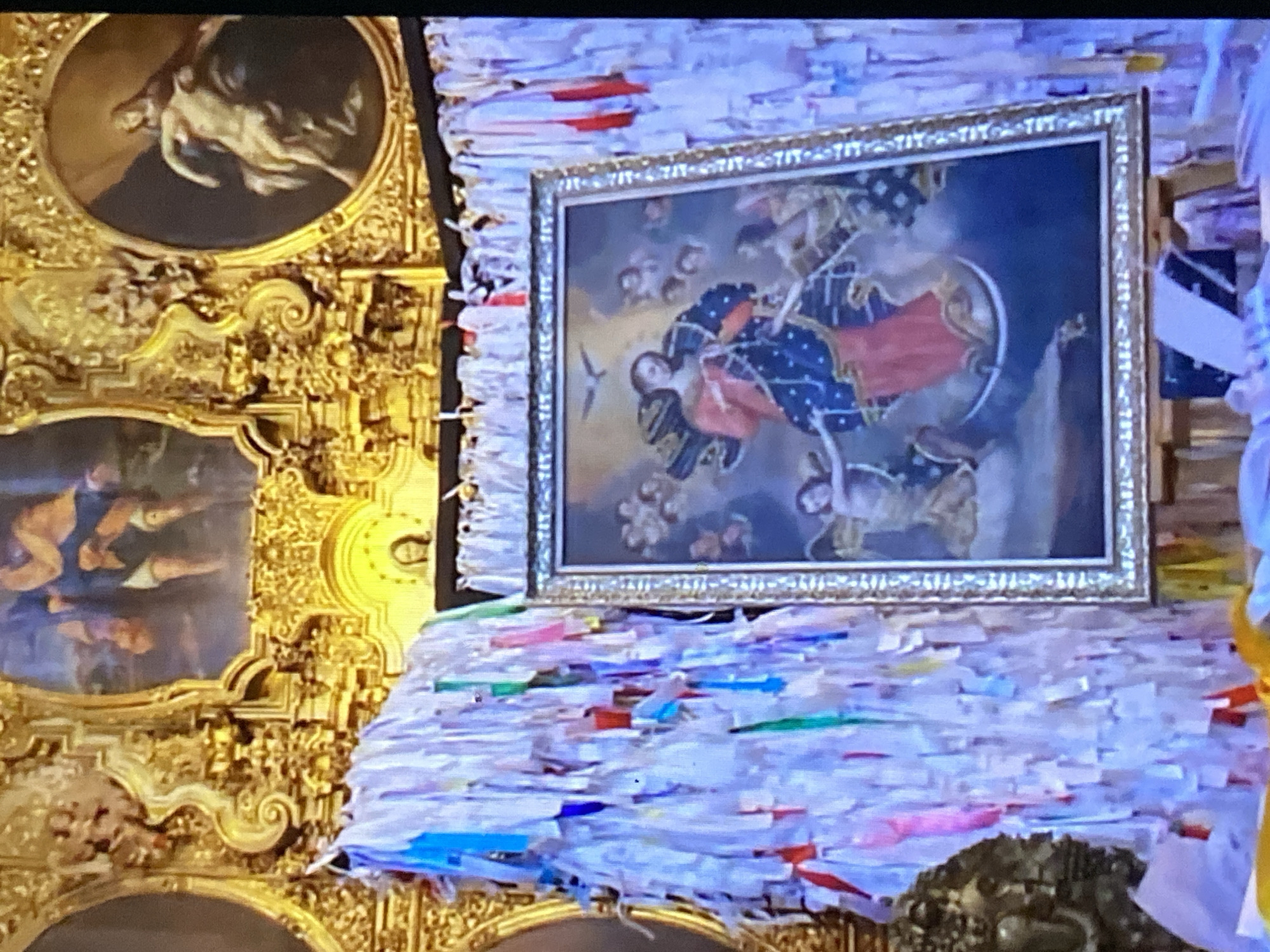 The image depicts the Altar of the Virgin of the Miraculous Medal in Tinaquillo, Venezuela. A rectangular framed painting of the Virgin Mary, in red and blue robes, surrounded by angels, is centered. Underneath the painting is a pile of small white papers with colored strips. To the left are two circular and square-shaped ornate gold framed images, possibly other paintings or sculptures. The entire scene is a display or shrine.