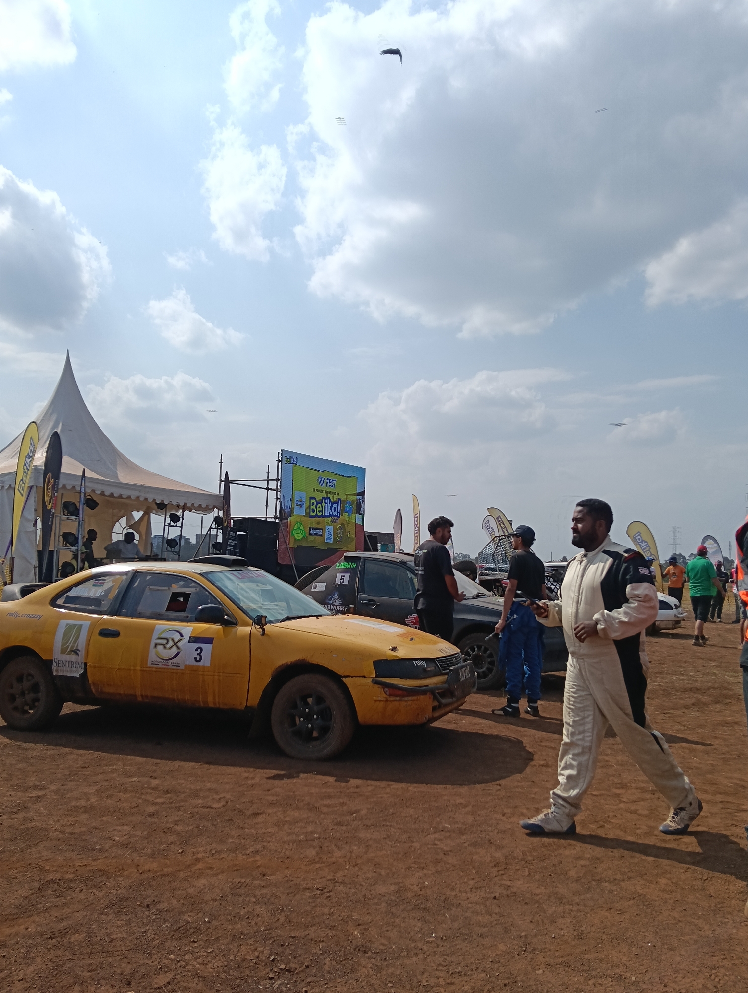 A man in a white and black racing suit walks across a dirt track towards the right of the frame. He is looking to his right. Behind him, a yellow rally car with a "3" on its door is parked. To the left of the car is a white tent with yellow banners. In the background, a large screen displays "Betika! RX FEST". Other people and cars are visible in the distance, as well as some power lines. The scene appears to be an outdoor event, possibly a race, under a bright, cloudy sky.
