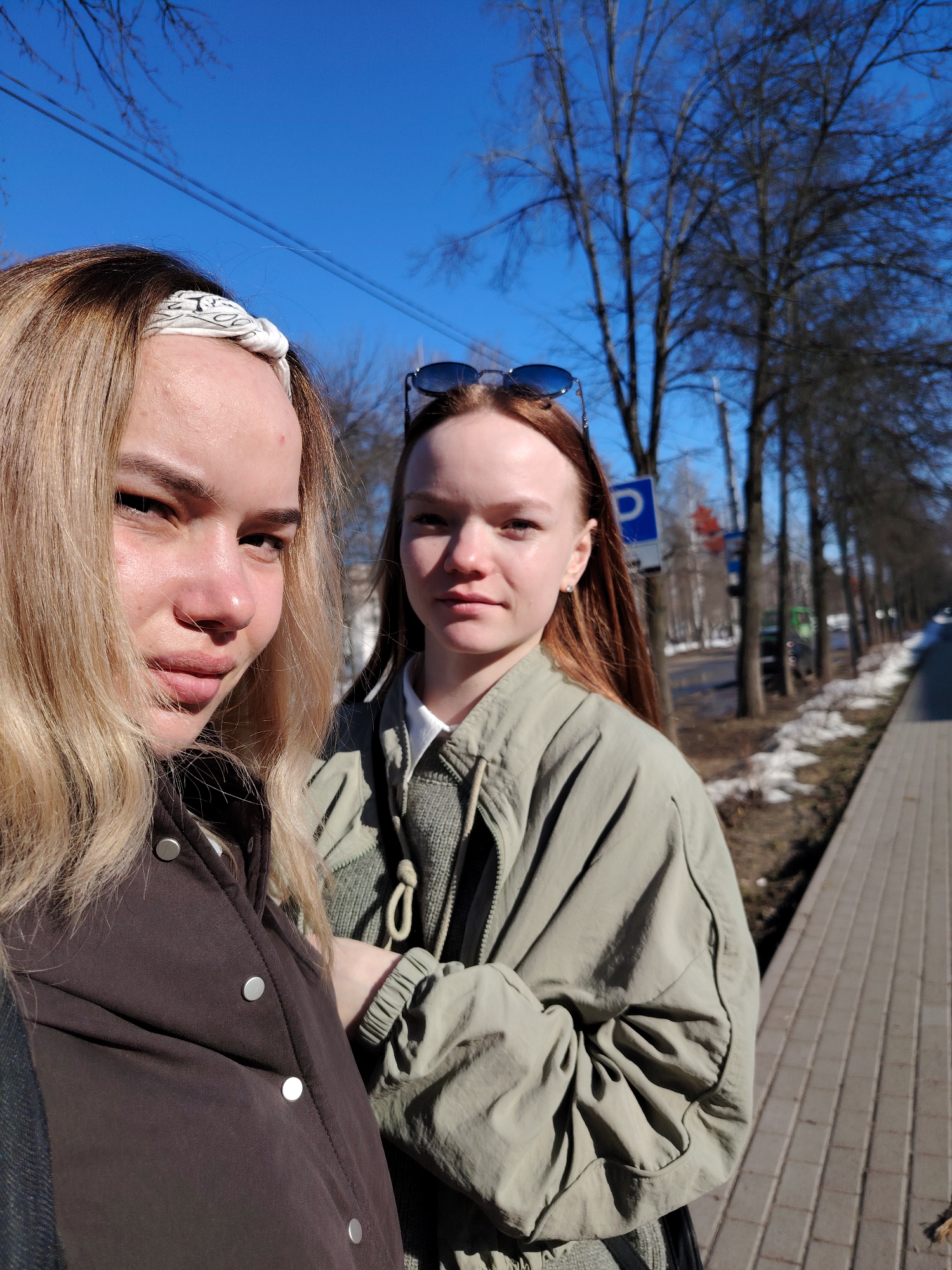 Two young women are standing on a paved sidewalk. The woman on the left has blond hair and is wearing a dark brown jacket with silver buttons and a patterned white headband. The woman on the right has auburn hair and is wearing a light green jacket with sunglasses perched on her head. Behind them is a street with bare trees, a blue sky, and remnants of snow along the roadside. A blue parking sign is visible in the background. The location is Chaikovsky, Russia.
