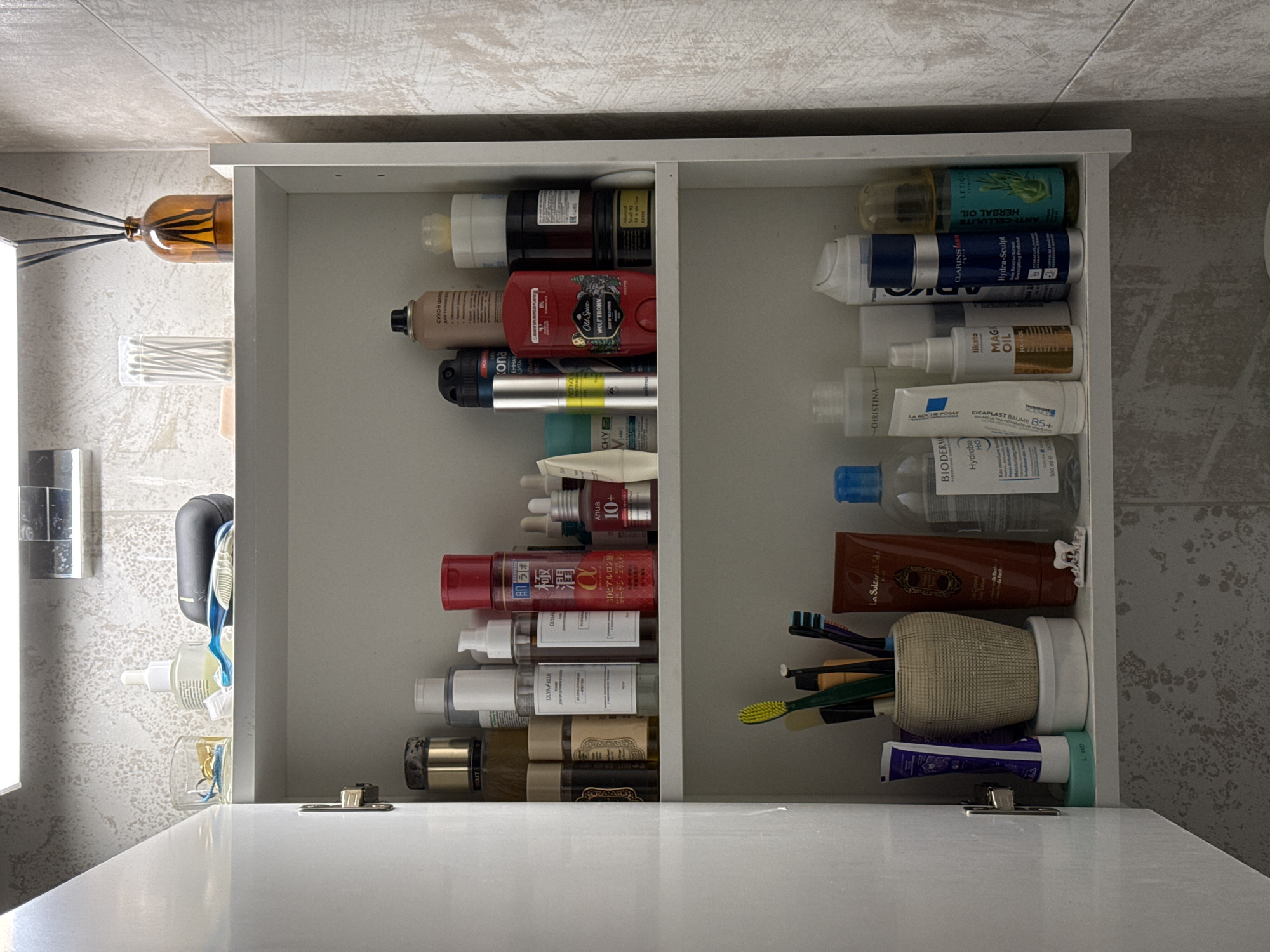 This is a top-down view of an open white bathroom cabinet, meticulously organized and packed with various personal care and cosmetic products. The cabinet is divided into two main vertical sections, each with multiple compartments.
On the left side of the cabinet, outside of it, a brown glass reed diffuser with black reeds is visible, along with a container of cotton swabs, a dark grey electric shaver or speaker, and a clear glass holding toothbrushes and a small brush. The background features light grey, textured wall tiles.
Inside the left section of the cabinet, numerous products are stored horizontally. Visible items include a clear bottle of LETIQUE ANTI-CELLULITE HERBAL OIL, a dark jar labeled "Snail 92," a can of KONA dry shampoo with Cyrillic text, a red bottle of Old Spice WOLFTHORN, and a silver tube of CHRISTINA RETINOL. Further down, there are products with Cyrillic labels such as "DLYA KOJI," a red bottle of Hada Labo Gokujyun with Japanese text (3Dヒアルロン酸・コリーダシーエラスチン), an Anua 10+ bottle, and a brown tube from La Sultane de Saba. A golden bottle of Likato MAGI OIL for Hair Care is also visible.
The right section of the cabinet holds more products, including a blue and white spray bottle of CLARINS Hydra-Sculpt, a white tube of LA ROCHE-POSAY CICAPLAST BAUME B5+, and a large BIODERMA Hydrabio H2O bottle. The bottom compartment of this section contains a brown tube of La Sultane de Saba, a beige cup holding several toothbrushes (blue, green, yellow), and a purple and white tube of toothpaste.
The presence of Cyrillic text on products like KONA dry shampoo and labels such as "DLYA KOJI" (for skin) are strong indicators supporting the Moscow, Russia location context. The scene is brightly lit, suggesting indoor lighting. There are no people visible, and no specific activity is taking place other than the display of stored items.